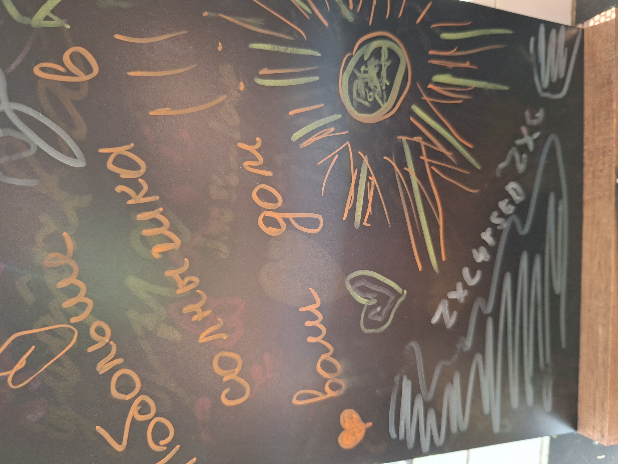 The image is a close-up, angled shot of a chalkboard with chalk drawings and text. The chalk is in various colors, including orange, yellow, green, and white. The writing appears to be in Russian. The visible text translates to "больше солнца ваш дом" which means "more sun your home." There are also drawings of a heart, a sun, and scribbled lines. The chalkboard is mounted on a wooden stand, suggesting it might be displayed in a public space or establishment. The lighting in the image is dim, with the chalk colors standing out against the dark background. The overall impression is of a warm, inviting message or decoration. There is no indication of the city or specific location.