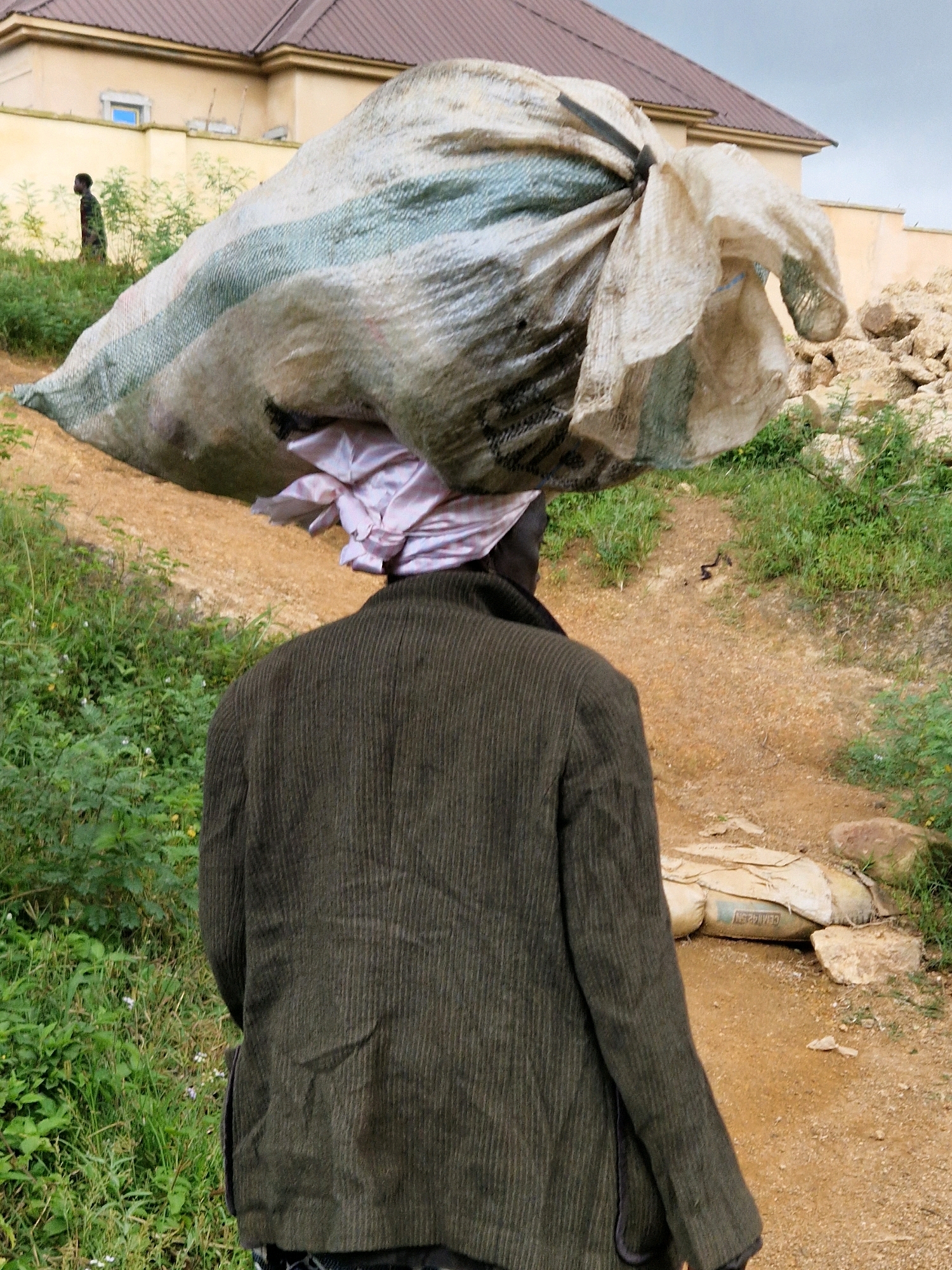 A person, seen from behind, walks along a dirt path carrying a large, white and green striped sack of goods on their head, supported by a folded pink fabric. The person is wearing a dark green corduroy jacket. The environment is outdoors with green foliage and dirt on either side of the path. In the background, a tan-colored building with a brown roof is visible, as well as a pile of rocks and some construction materials. A small, dark figure can be seen walking in the distance to the left.
