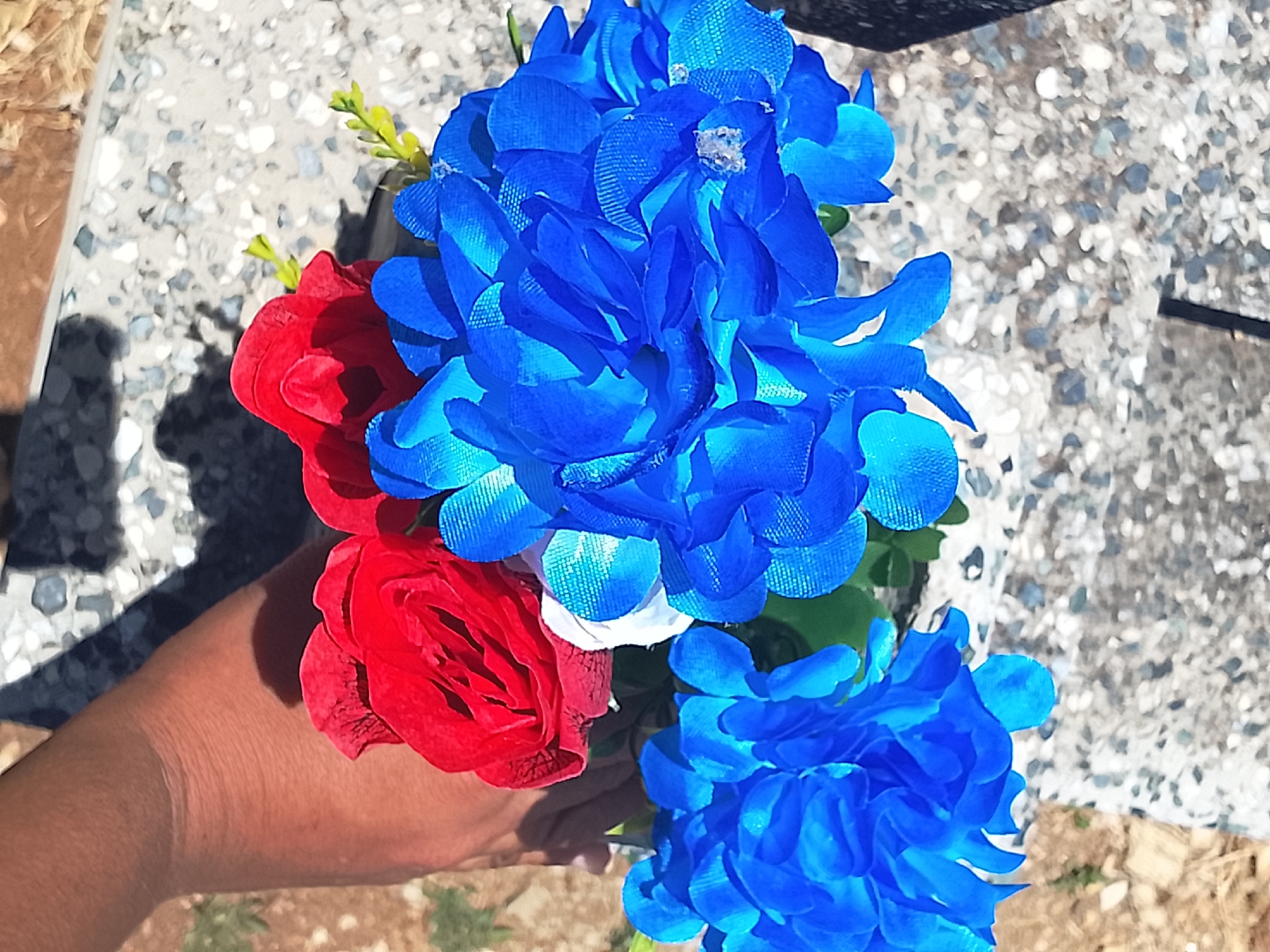 A hand holds a bouquet of artificial red, white, and blue flowers. The background consists of a mottled gray and white surface. The location is Altagracia, Venezuela.
