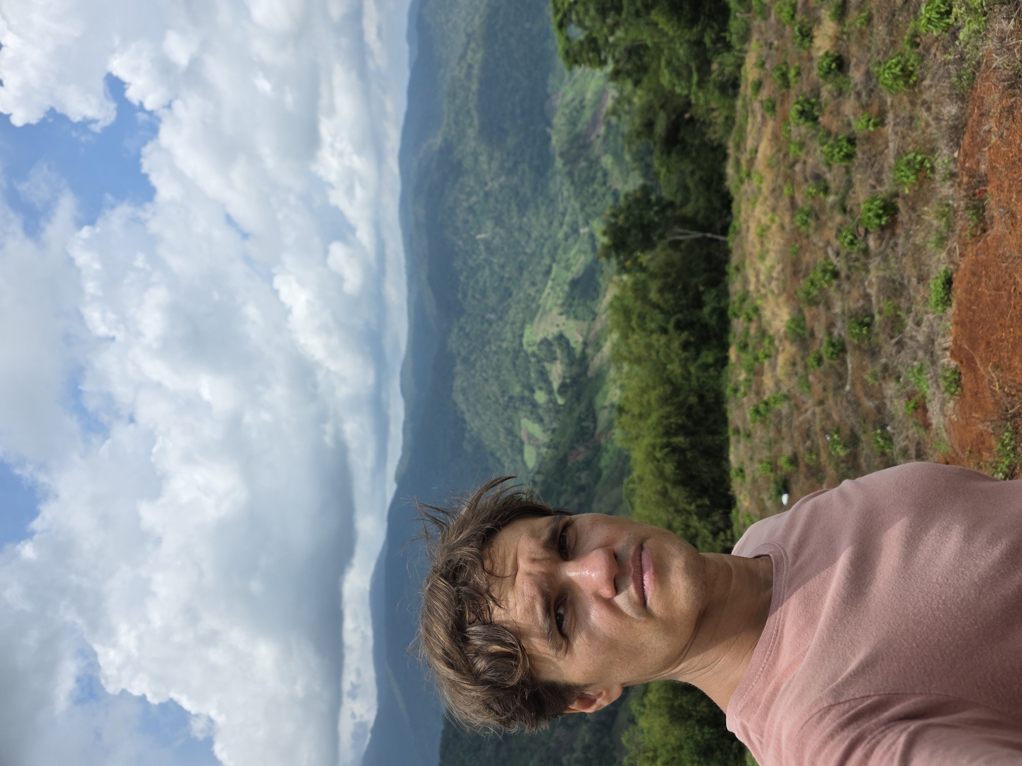 A man takes a selfie in front of a lush, mountainous landscape in Nao Sẻ, Vietnam. The man, who appears to be in his late 30s or early 40s, is wearing a pink t-shirt and has messy brown hair. His expression is serious as he looks directly at the camera.
The background is a panoramic view of rolling hills covered in dense green vegetation. Terraced fields are visible on some of the slopes, indicating agricultural activity. The sky above is a mix of blue patches and large, fluffy white clouds. In the foreground, there is a patch of dry, reddish-brown soil with scattered green bushes and some dry grass. Behind the man, a line of green trees forms a natural border. The overall impression is one of natural beauty and a serene, remote location.