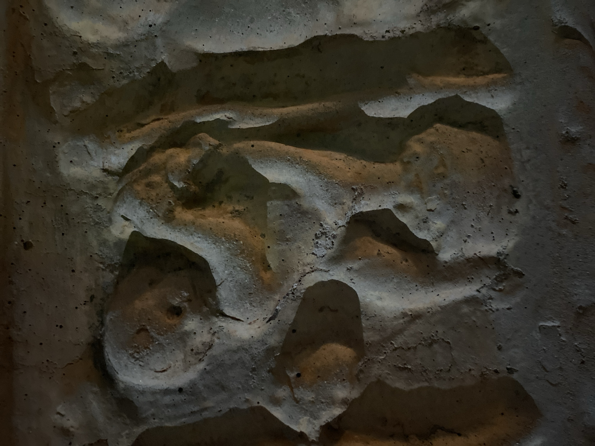 This image depicts a section of a rough, textured wall. The material appears to be stone or concrete with uneven surfaces and varying shades of gray, beige, and brown. The lighting is dim, creating shadows that accentuate the irregular shapes and crevices of the wall. There are no people, animals, or identifiable objects within the frame other than the wall itself. There is insufficient information to place the image at a specific location in Budva, Montenegro.