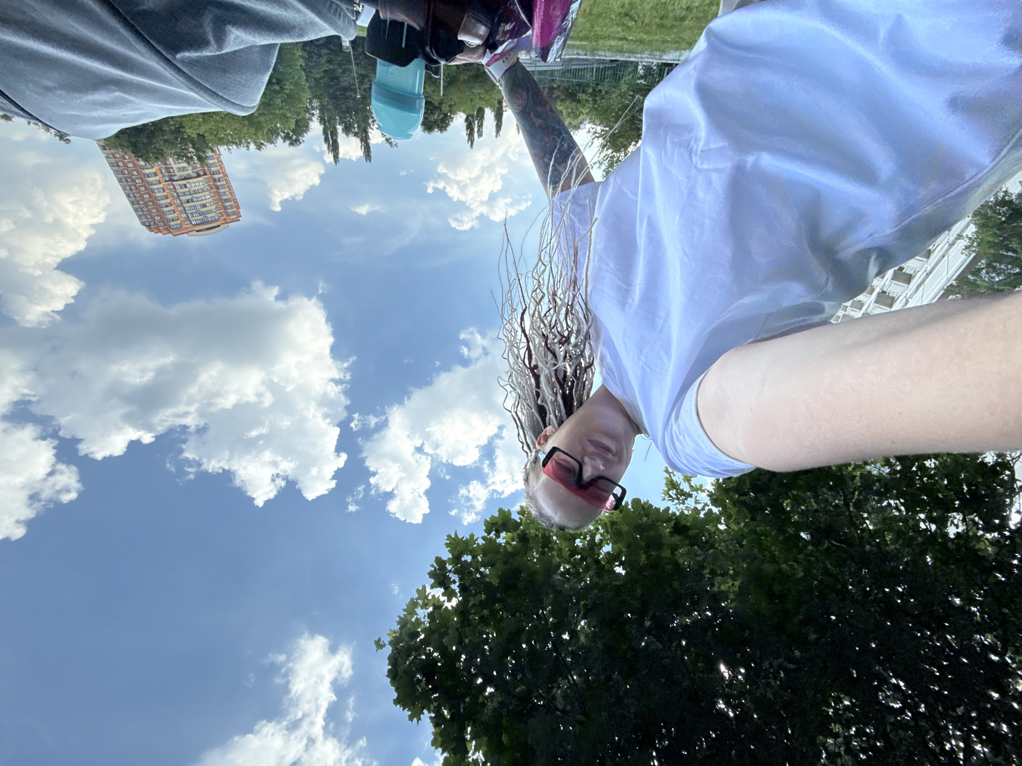 An extreme low-angle outdoor selfie captures a person with blonde dreadlocks and pink-rimmed sunglasses. They are wearing a white t-shirt. Their left arm is visible with a tattoo. In the background, lush green trees are abundant, with a modern, orange-brick building visible in the distance under a bright blue sky with scattered white clouds. The location is identified as Moscow, Russia.
