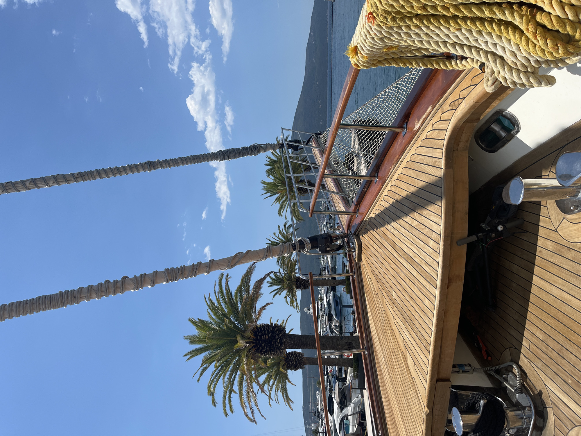 The image captures a close-up, forward-facing view from the deck of a wooden boat, likely at the Porto Montenegro marina in Tivat, Montenegro. The boat's polished wooden deck takes up the foreground, with metal cleats and coiled ropes visible. Two thick, textured ropes are prominently featured, one hanging over the railing on the right side of the frame, appearing weathered and yellowed.
Beyond the boat's railing, a marina filled with various yachts and smaller boats is visible. Several palm trees, characteristic of the Mediterranean climate, stand tall between the boats and the water. In the background, a lush, green mountain range rises from the coastline, under a clear blue sky dotted with scattered white clouds. The water appears calm and blue, reflecting the bright daylight. The scene evokes a sense of a warm, sunny day in a luxurious maritime setting.