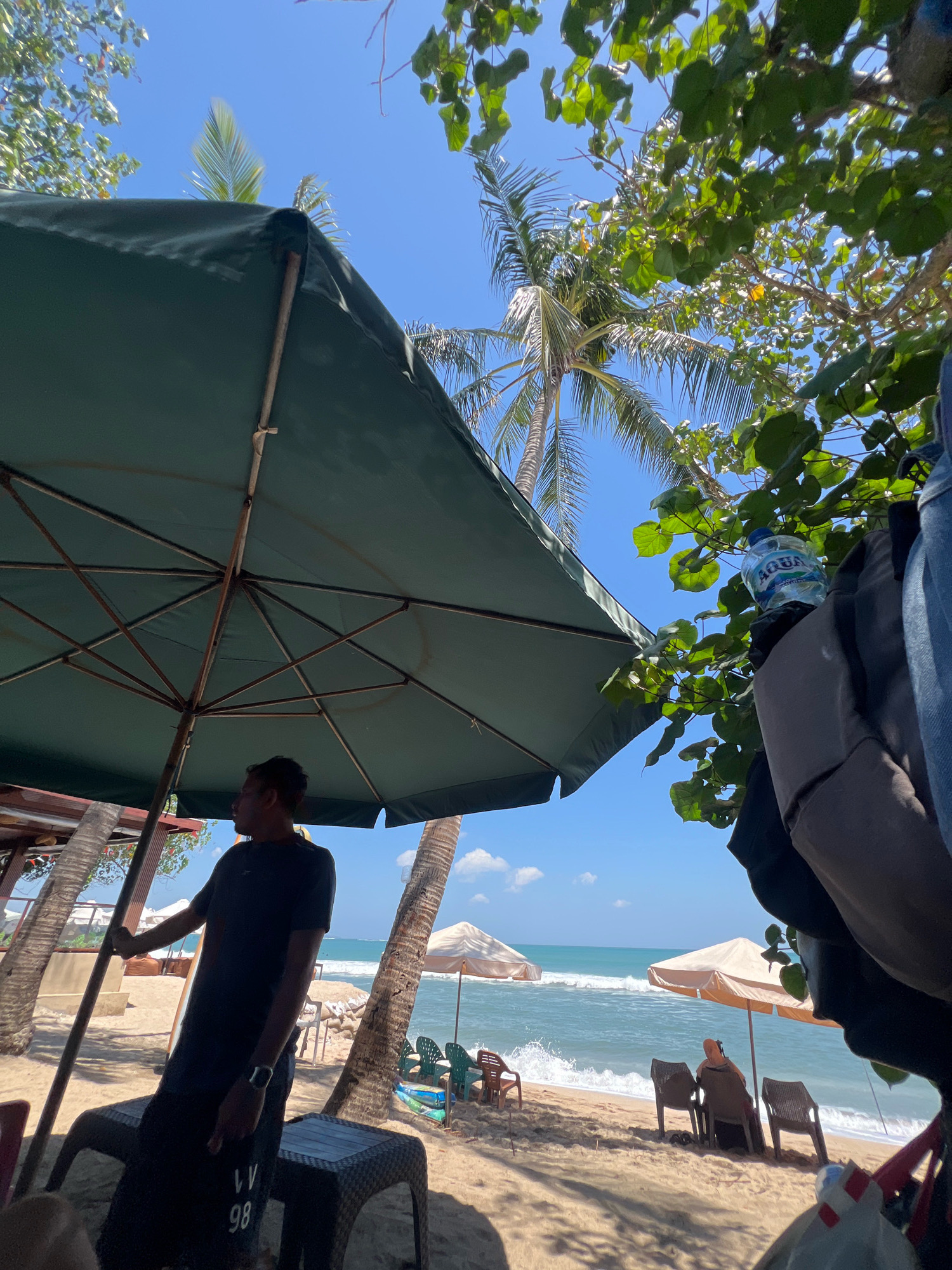 The image depicts a sunny beach scene in Kuta, Indonesia, likely during the daytime. A large, dark green beach umbrella dominates the foreground, casting shade. A man, wearing a dark t-shirt and shorts with the number "98" printed on them, stands under the umbrella, looking out towards the ocean. He is wearing a wristwatch. In the background, the turquoise ocean meets a sandy beach, with gentle waves crashing ashore. Several beach umbrellas, some lighter in color, are scattered along the beach, with chairs positioned nearby. Palm trees frame the scene, with lush green foliage from a tree in the upper right corner of the image. A plastic bottle of "AQUA" water is visible, placed on what appears to be a backpack or bag. The sky is a clear blue with some faint clouds. The overall atmosphere suggests a relaxed, tropical vacation setting.