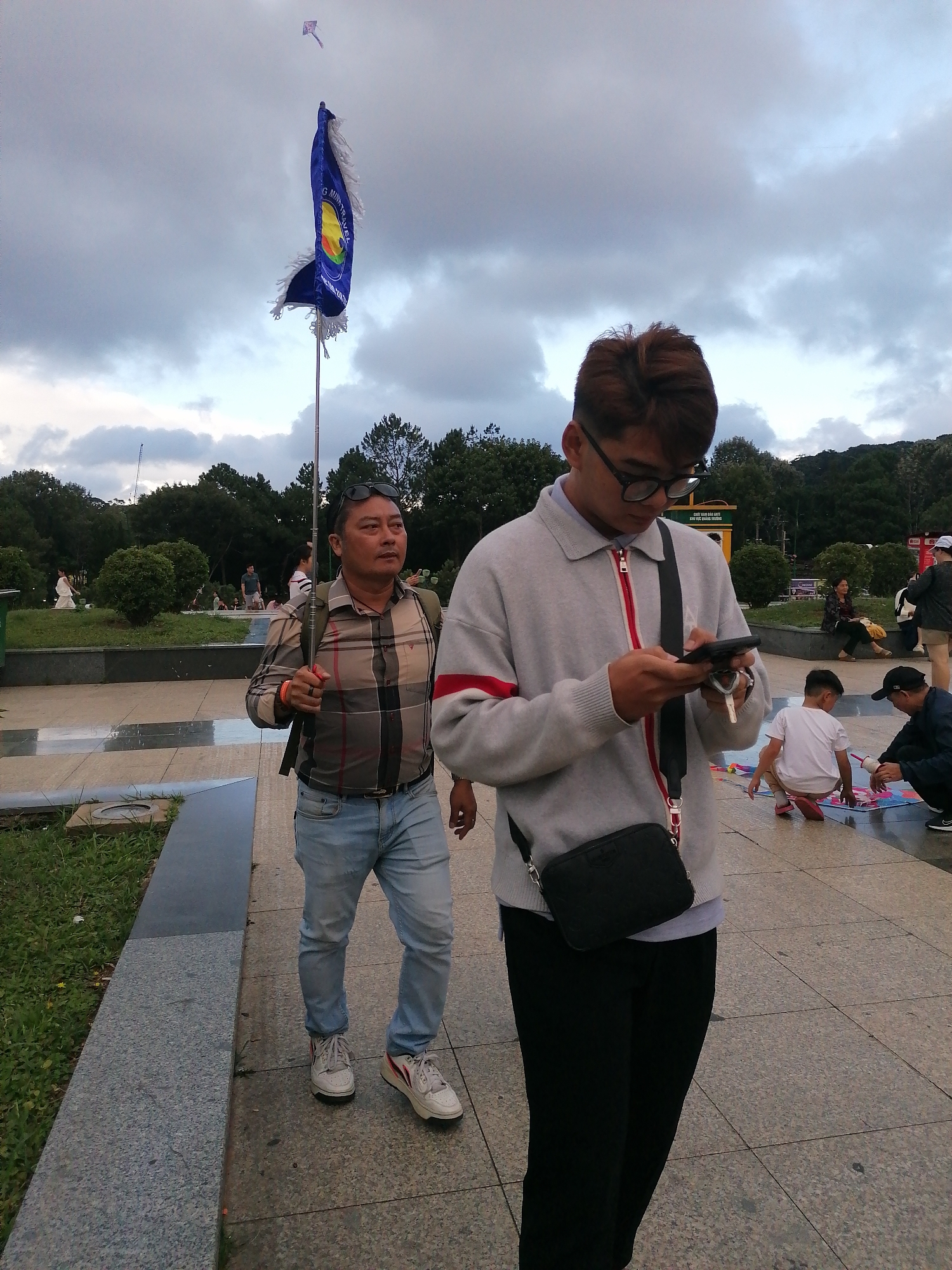 This image depicts a cloudy day in Da Lat, Vietnam, likely in an outdoor public space with paved walkways and greenery. In the foreground, a young man with short, styled brown hair and glasses is looking at his phone while walking. He is wearing a light grey zip-up sweater with red and white striped accents on the sleeves and collar, a black crossbody bag, and black pants. Behind him, a middle-aged man with sunglasses on his head and a plaid shirt is walking and holding a long pole. He is wearing light blue jeans and white sneakers, and a backpack. In the background, several other people are visible, including children playing with kites on the ground, and some individuals sitting or walking in the distance. A blue flag with white fringe is planted on a pole. The overall atmosphere appears to be relaxed and casual.