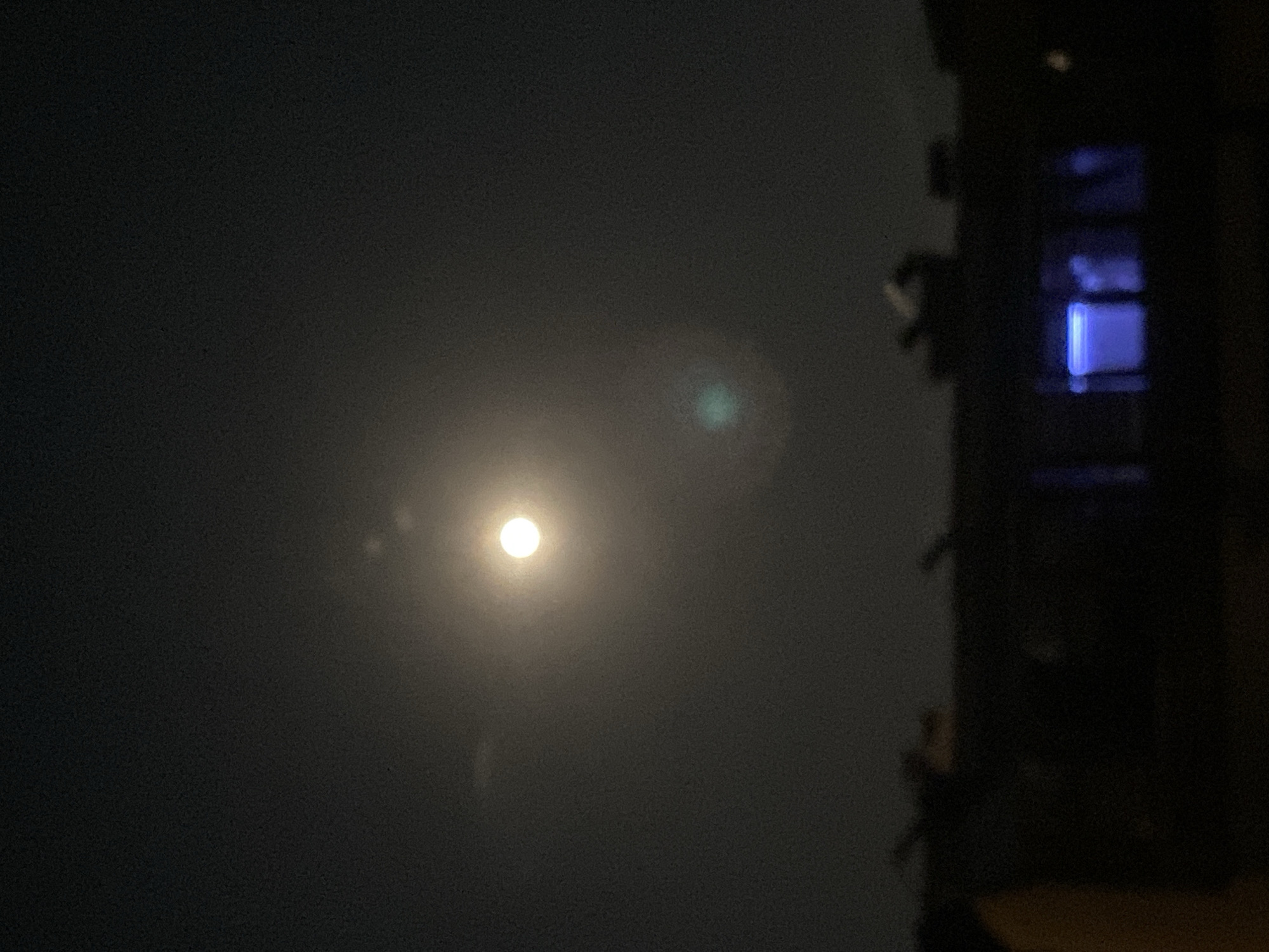 The image depicts a dark, possibly nighttime, scene in Varna, Bulgaria. The sky is overcast and dark. A bright light source, likely the moon or a strong street lamp, is visible through the haze in the upper center of the frame. To the right, a dark, silhouetted structure suggests a building or part of a building with visible windows, and a blue light emanates from one of the windows. The surroundings are generally obscured by darkness, suggesting a low-light environment.