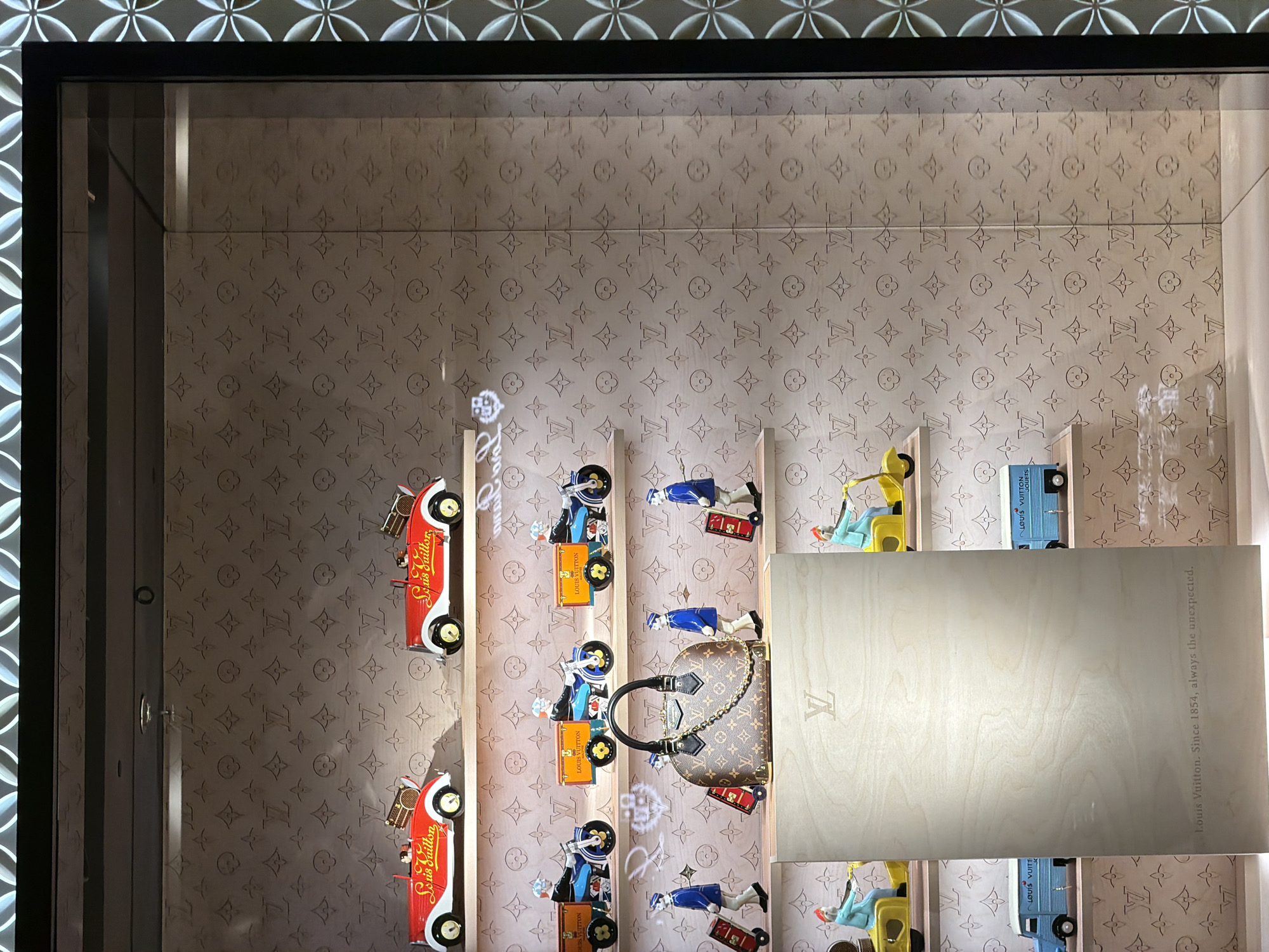 The image depicts a retail display window, likely within a Louis Vuitton store in Xuanwu District, China. The display is organized into multiple shelves. The background of each shelf features a repetitive pattern of the Louis Vuitton monogram.
The top shelf showcases two red toy cars with white accents, each carrying a miniature brown Louis Vuitton trunk on its rear. The second shelf from the top displays three toy motorcycles, each ridden by a miniature figure dressed in a blue uniform and cap, pulling a yellow cargo box.
The third shelf features a central, medium-sized Louis Vuitton handbag with a prominent gold chain strap. Flanking the handbag are four miniature figures dressed in blue uniforms and caps, each interacting with small, red rolling suitcases.
The fourth shelf from the top has two yellow toy scooters, each ridden by a miniature figure wearing a blue uniform and red hat.
The bottom shelf is dominated by a large, light-colored wooden panel, seemingly a display stand. A Louis Vuitton logo is embossed near the top of this panel, and text at the bottom reads "Louis Vuitton. Since 1854, always the unexpected." On either side of this wooden panel, at the very bottom of the visible display, are two light blue toy vans, each with "LOUIS VUITTON JOUETS" printed on its side.