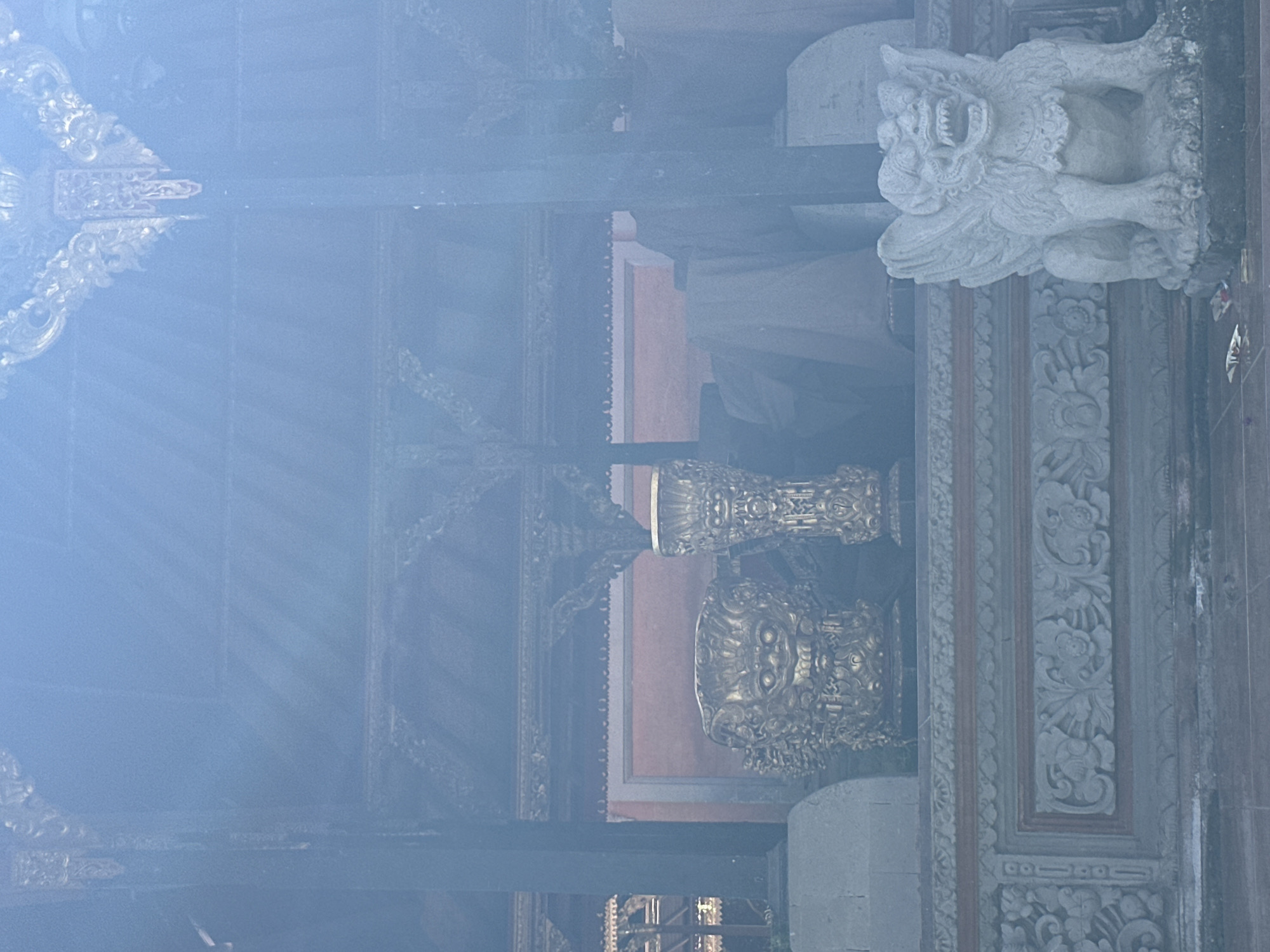 A stone statue of a mythical creature with wings and an open mouth is situated to the right of a tiered stone platform in what appears to be an outdoor temple or shrine. The platform is decorated with intricate carvings and a red border. To the left of the platform, two ornate golden vases or urns are placed. One of the golden urns is a large, elaborate piece shaped like a monstrous face. Behind the golden objects, fabric is draped over unseen objects, and a red wall is visible. The scene is bathed in a bright, hazy light, suggesting it may be a sunny day or that there is a light haze in the atmosphere. The background features architectural elements with decorative wooden carvings and dark pillars, characteristic of Balinese architecture, possibly in Ubud District, Indonesia.