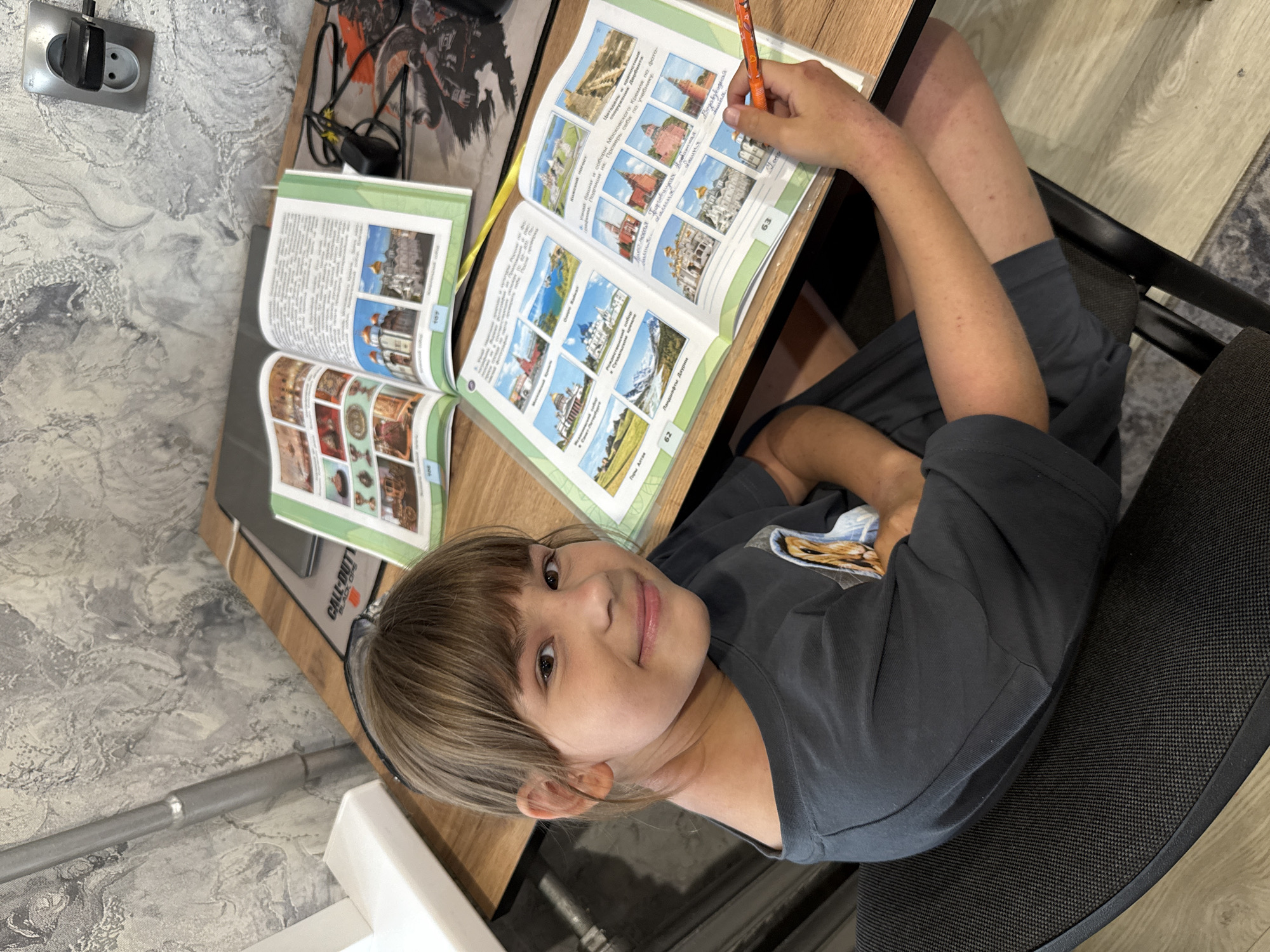 A young person is seated at a desk and working in a book. The book is open to pages that show architectural landmarks and geographical features, with handwritten labels. The child is holding a pencil, and appears to be writing in the book. The desk also has another book open, a mousepad with a "Call of Duty" logo, and a laptop. The location is inside a room, with a patterned wall visible. There are no indications of Rusanovo.