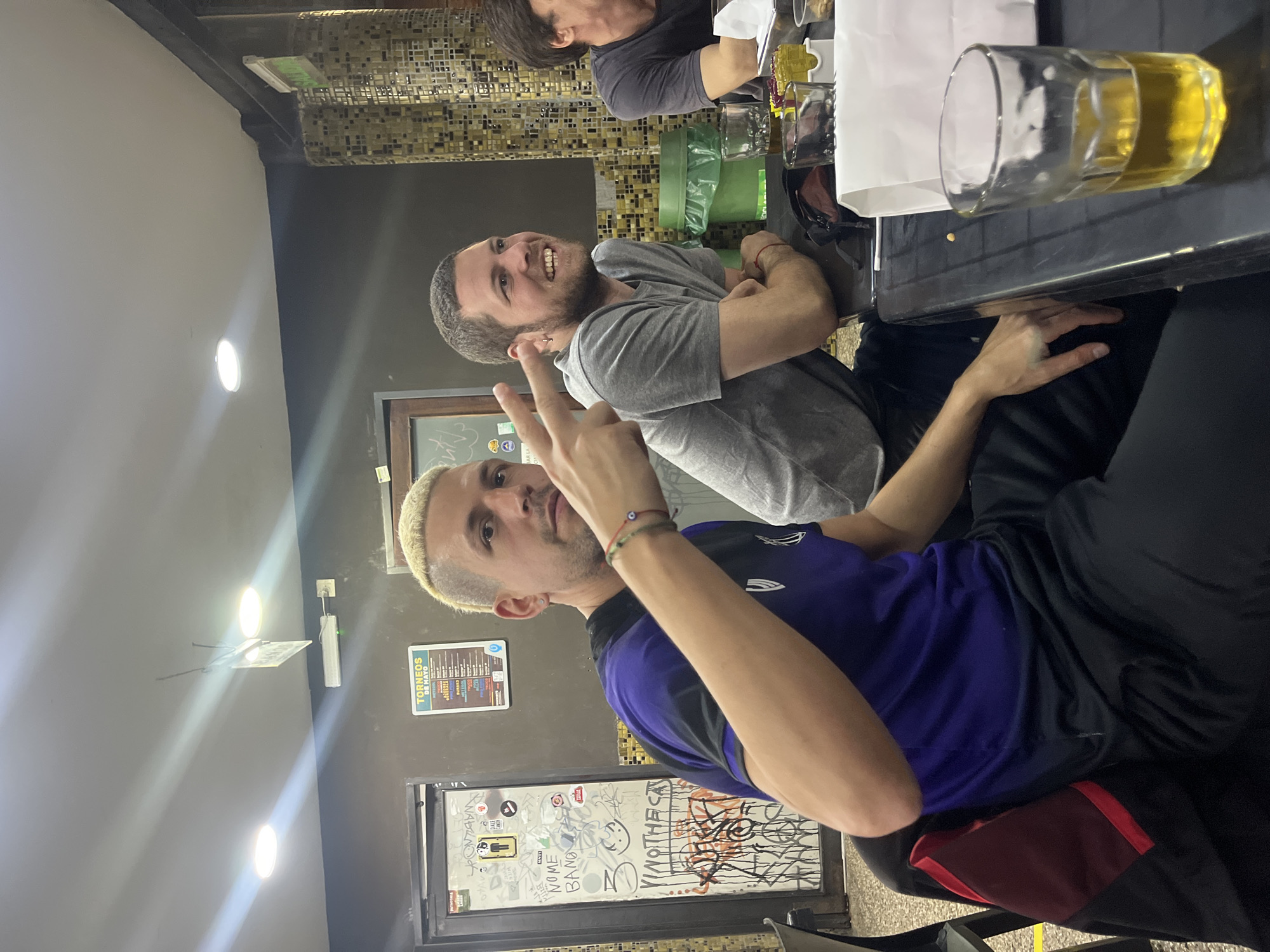 The image shows three people at a bar. Two men are facing the camera, while the third person is only partially visible in the background. The first man is making a peace sign, wearing a blue and black shirt, and has bleached hair. The second man is wearing a grey shirt with his arms crossed and is smiling at the camera. There are beer glasses on the table, and there are graffiti-covered walls and a menu behind the men. Based on the menu visible on the wall listing "Torneos de Mayo" and the overall aesthetic, the location is likely a bar in Buenos Aires, Argentina.