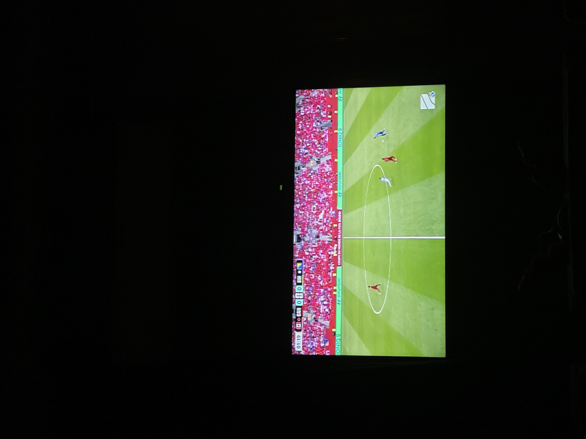 The image shows a television screen displaying a soccer match. The score on the screen indicates CAN 0 - 0 BIH, with the time at 01:19. The stadium is filled with spectators wearing red. A promotional banner with "HYUNDAI" and "IONIQ 9" is visible at the bottom of the screen, along with "COUPE DU MONDE DE LA FIFA 2026". Two players are on the green field, one in a red uniform and the other in a blue uniform, with a soccer ball between them. The television is in a dark room, suggesting it is nighttime or the room is intentionally kept dark for viewing. There are hints of people or furniture in the foreground, but they are largely obscured by darkness. The location is identified as Tbilisi, Georgia.