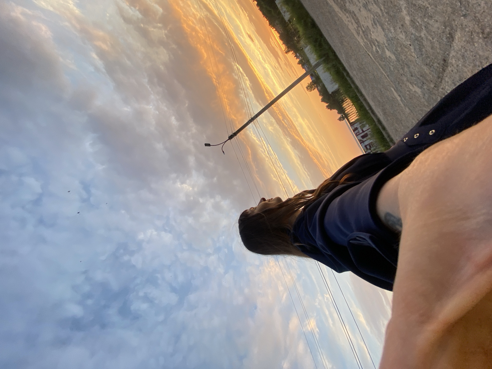A low-angle, wide shot captures a young woman standing outdoors during sunset. She is seen from the chest up on the right side of the frame, looking off to the right. She has long, dark brown hair and is wearing a dark blue hooded jacket. Her left arm is extended towards the camera, with her hand and forearm prominently in the foreground, obscuring part of her body.
The background features a dynamic sky filled with clouds, transitioning from pale blue at the top to vibrant oranges and yellows near the horizon, indicating the setting sun. A utility pole with a light fixture stands tall on the right side of the frame, with power lines stretching across the sky. In the distance, there are trees, buildings, and a body of water. The ground in the foreground is a paved surface. The scene appears to be in Votkinsk, Russia, based on the overall atmosphere and landscape elements.
