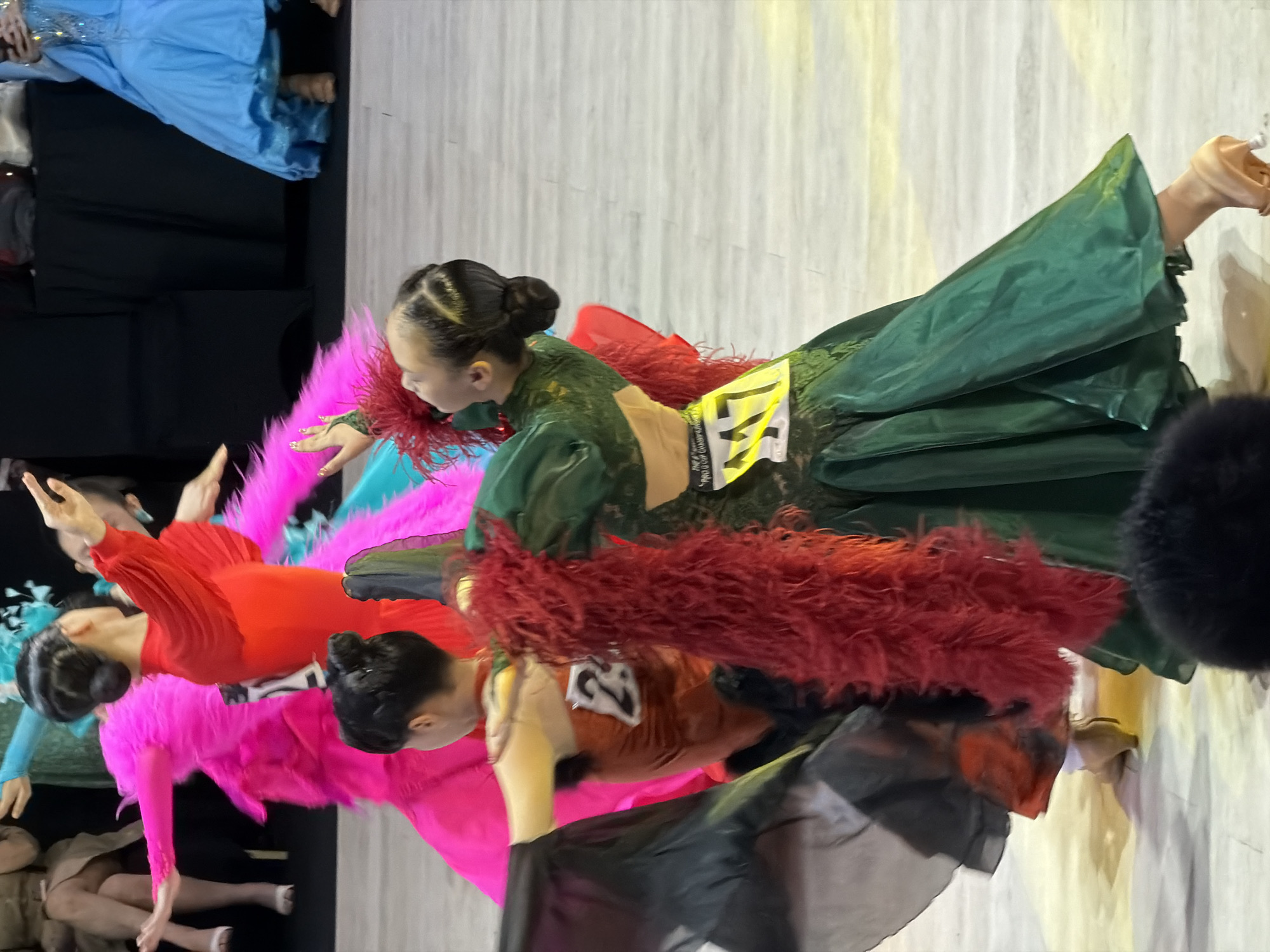 The image captures a scene of ballroom dancing competition, likely in Hanoi, Vietnam, given the context of similar events held there. Two young female dancers are the central focus. The dancer in the foreground is wearing a vibrant green ballroom dress with intricate detailing and a feather boa around her neck. Her hair is styled in an updo. She is in motion, her arms extended, and her skirt is flowing. She has a competition bib with the number "M7" on it. To her left, another dancer, possibly her partner or a competitor, is seen in an orange costume with a bib displaying the number "23". Her back is partially turned towards the viewer. In the background, other dancers and audience members are visible, including a person in a bright pink costume and another in a blue dress. The floor appears to be a polished surface, possibly a ballroom floor or stage. The lighting suggests an indoor event with spotlights illuminating the dancers. The overall atmosphere is one of energetic performance and competition.