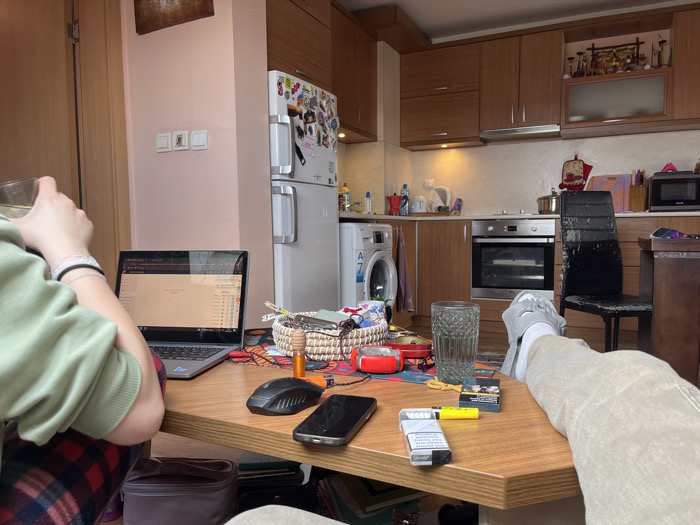 The image displays an interior scene, identified as being located in Sofia, Bulgaria, combining a kitchen and a casual living area. Two individuals are partially visible. On the left, a person wearing a light green long-sleeved top and red and black plaid pajama bottoms holds a clear, textured glass in their right hand. An open silver laptop rests on a hexagonal wooden table directly in front of this individual, displaying a web interface with a list and content. On the right, a second individual's legs, clothed in light-colored trousers and grey slippers, are extended onto the same wooden table.
The table's surface contains a second clear, textured glass, a black computer mouse, a black smartphone placed screen-down, an open pack of white and blue cigarettes, a yellow lighter, and a closed dark blue cigarette pack. A woven basket holding various small items and a colorful placemat are also present on the table.
The background reveals a kitchen area with light brown wooden upper and lower cabinets. A white, double-door refrigerator covered with magnets and stickers stands against a light pink wall. To the left of a built-in silver oven and stovetop is a white front-loading washing machine (with "A 7" visible). The kitchen counters feature a white tiled backsplash, under-cabinet lighting, a red moka pot, other cooking utensils, a microwave oven, and a black chair with distressed upholstery. Various kitchen appliances and decorative elements are scattered across the counters and shelves.
