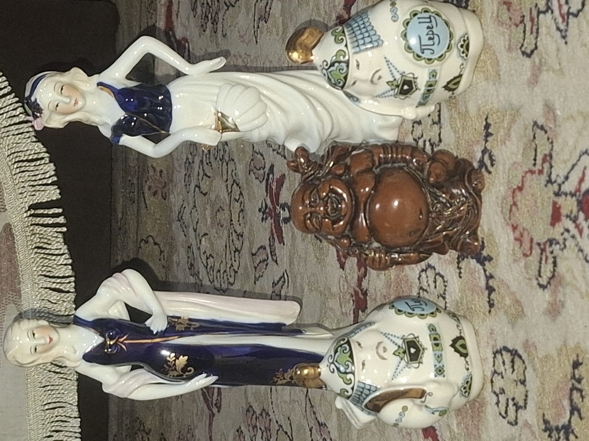 The image shows a collection of figurines arranged on a patterned carpet. On the left is a porcelain figurine of a woman in a long blue and white dress with gold accents, draped with a white shawl. To her right stands another porcelain figurine of a woman in a white dress with a blue bodice, holding a golden object. In the foreground, to the left, is a small, round, white ceramic pot with a decorative face and blue and green designs, possibly a kettle. In the center is a small, brown, ceramic figurine of a laughing Buddha, with his hands resting on his stomach. To the right of the Buddha is another small, round, white ceramic pot, similar to the one on the left, decorated with blue and green patterns and a blue oval with text. The background is blurred, showing a fringed textile and the continuation of the carpet pattern. The scene is static and appears to be an indoor display of decorative items. There is no indication of the city of Almaty, Kazakhstan, or any specific urban environment.