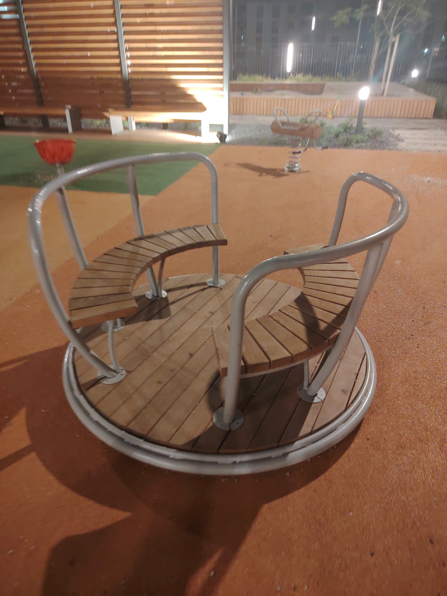 The image shows a merry-go-round with wooden seats and a metal frame. The merry-go-round is located on a brown, textured surface. In the background, there is a wooden structure with benches, a green area, and a fence. Several streetlights are visible, indicating it is nighttime. The scene appears to be a public park or playground. There are no people visible in the image.