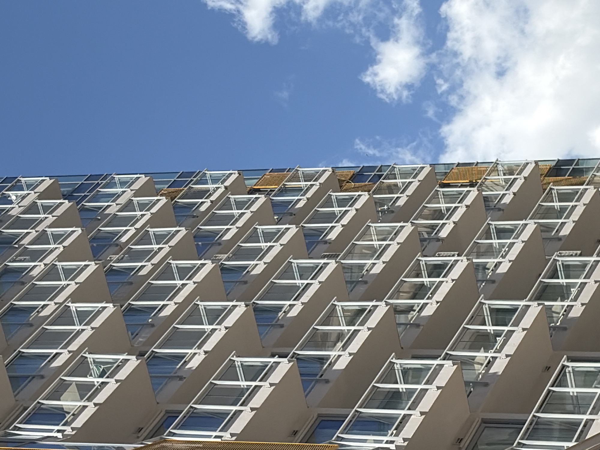 A high-angle, medium shot depicts the facade of a modern building in Rafailovići, Montenegro, against a blue sky with scattered white clouds. The building features a repeating geometric pattern of projecting, triangular structures with large windows. Some structures appear to have wooden balcony railings. No people are visible.