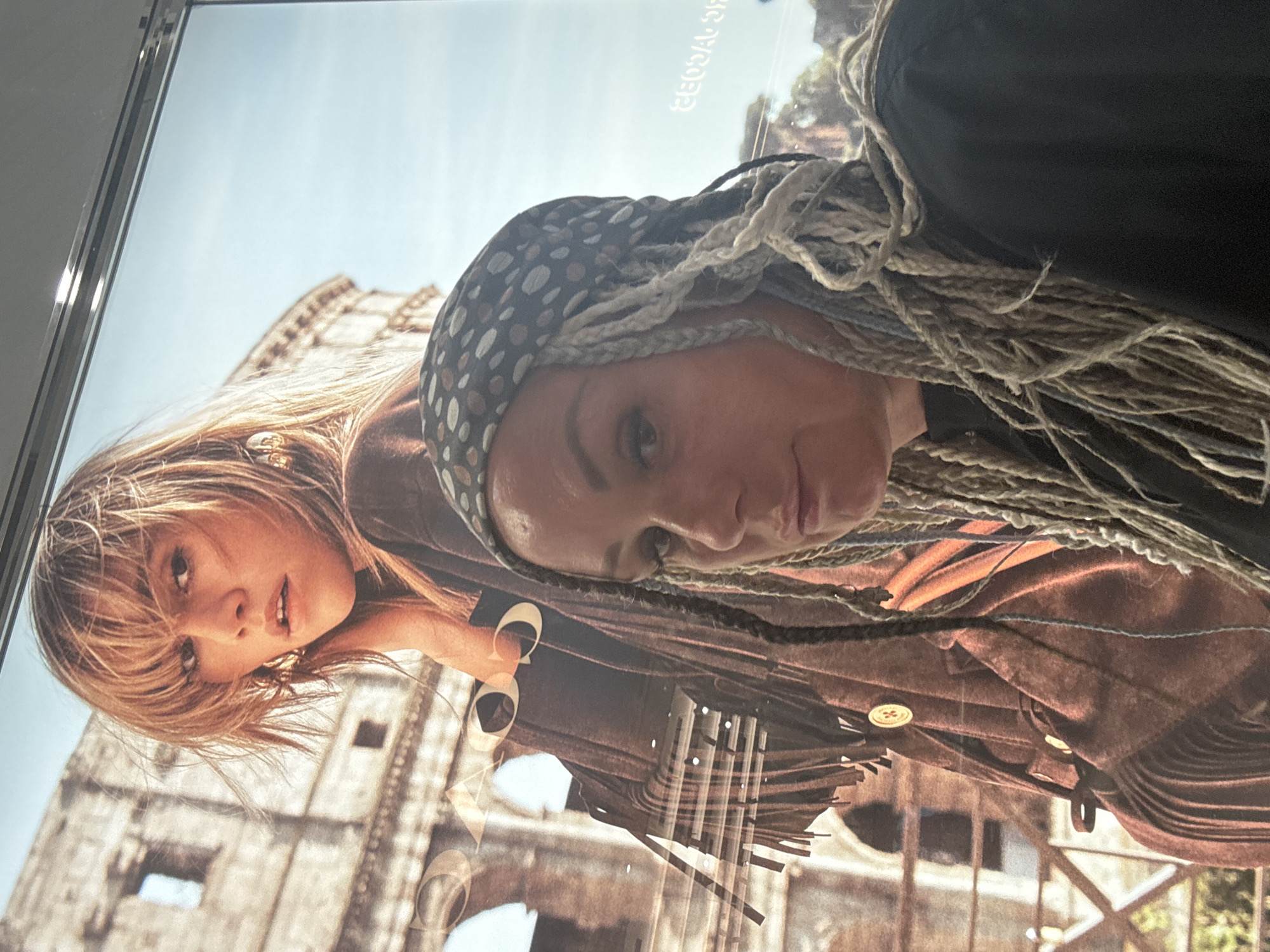 A woman with blonde hair is wearing a brown jacket and standing in front of the Colosseum in Rome, Italy. Another woman with braids and a scarf on her head is posing for the picture in front of the poster.
This photograph does not appear to be from Kuta, Indonesia.