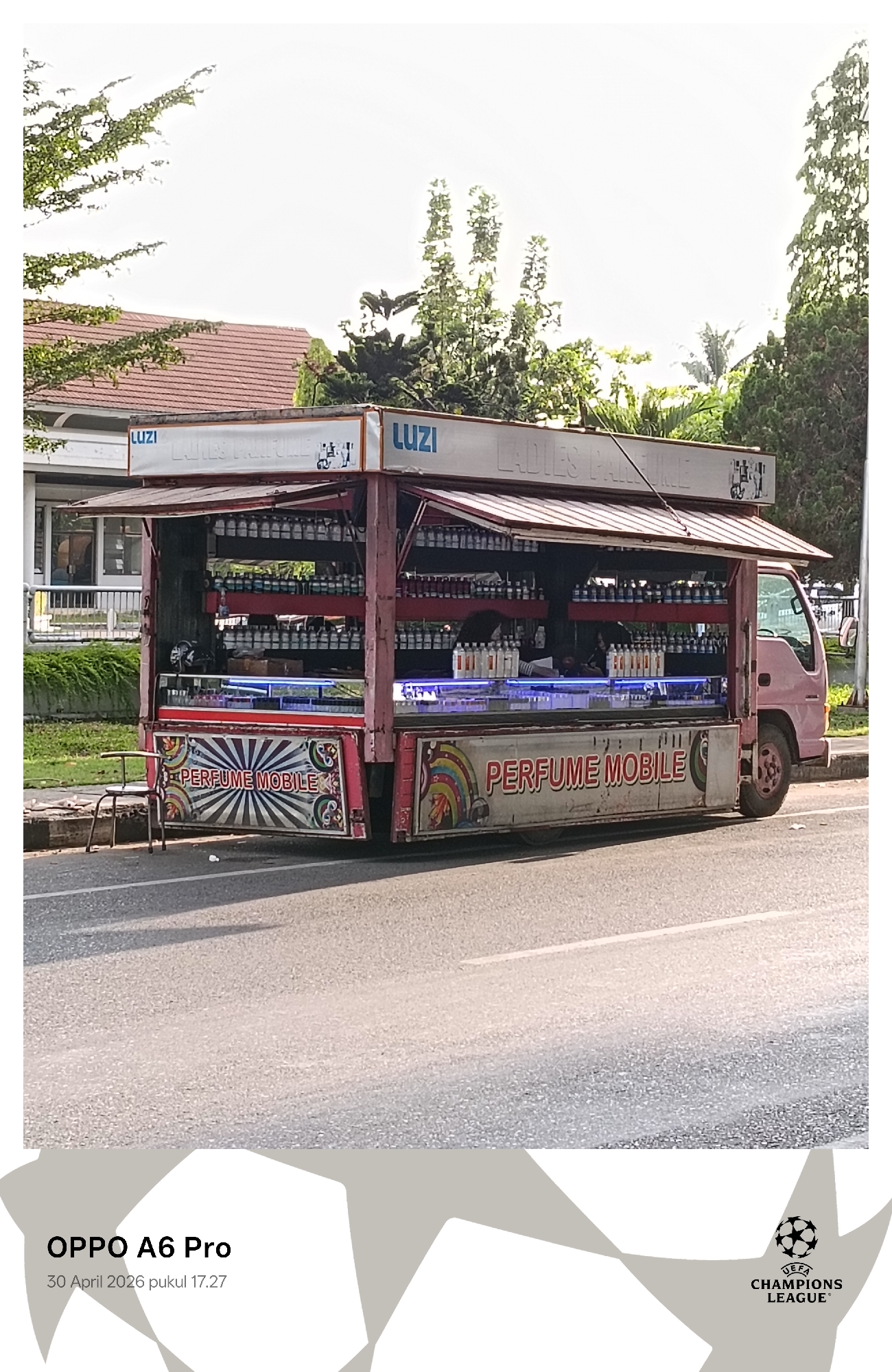 An outdoor, eye-level shot captures a vibrant pink commercial truck, converted into a mobile perfume shop, parked alongside an asphalt road. The truck features an open canopy revealing numerous shelves packed with hundreds of small, identical perfume bottles. A vendor is partially visible inside the truck, positioned behind a display counter that is illuminated by a blue LED strip. The side of the truck prominently displays the text "PERFUME MOBILE" in red letters, complemented by decorative patterns including a rainbow and starbursts. Banners on the truck's roof feature the text "LUZI" and the partially visible phrase "LADIES PERFUME."
To the left of the truck, a simple metal chair sits on the roadside. In the background, lush green trees and a white building with a red tile roof are visible. The bright, clear sky indicates late afternoon lighting, consistent with the image's timestamp. The scene depicts a typical roadside commercial setup, possibly in Lhokseumawe, Indonesia.
Visible text includes "LUZI," "LADIES PERFUME," and "PERFUME MOBILE." A watermark at the bottom left indicates "OPPO A6 Pro" and the capture date and time as "30 April 2026 pukul 17.27" (5:27 PM). A "UEFA CHAMPIONS LEAGUE" logo is also present as a watermark at the bottom right.