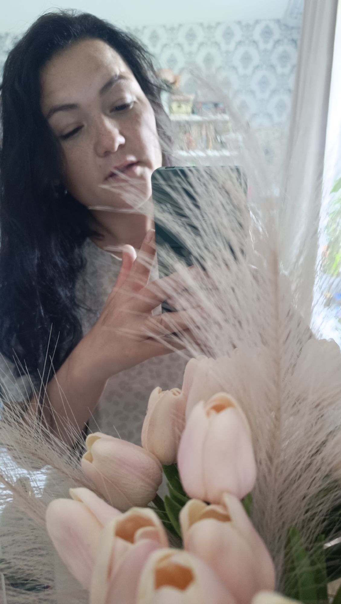 A woman with dark hair and freckles is looking down and holding a smartphone in her right hand, reflected in a mirror. She is wearing a light-colored, patterned shirt. In the foreground, a bouquet of light pink tulips and fluffy white plumes are visible. The background of the reflection shows a room with patterned wallpaper and shelves. The scene is likely indoors, possibly in a residential setting in Chaikovsky, Russia, given the context.