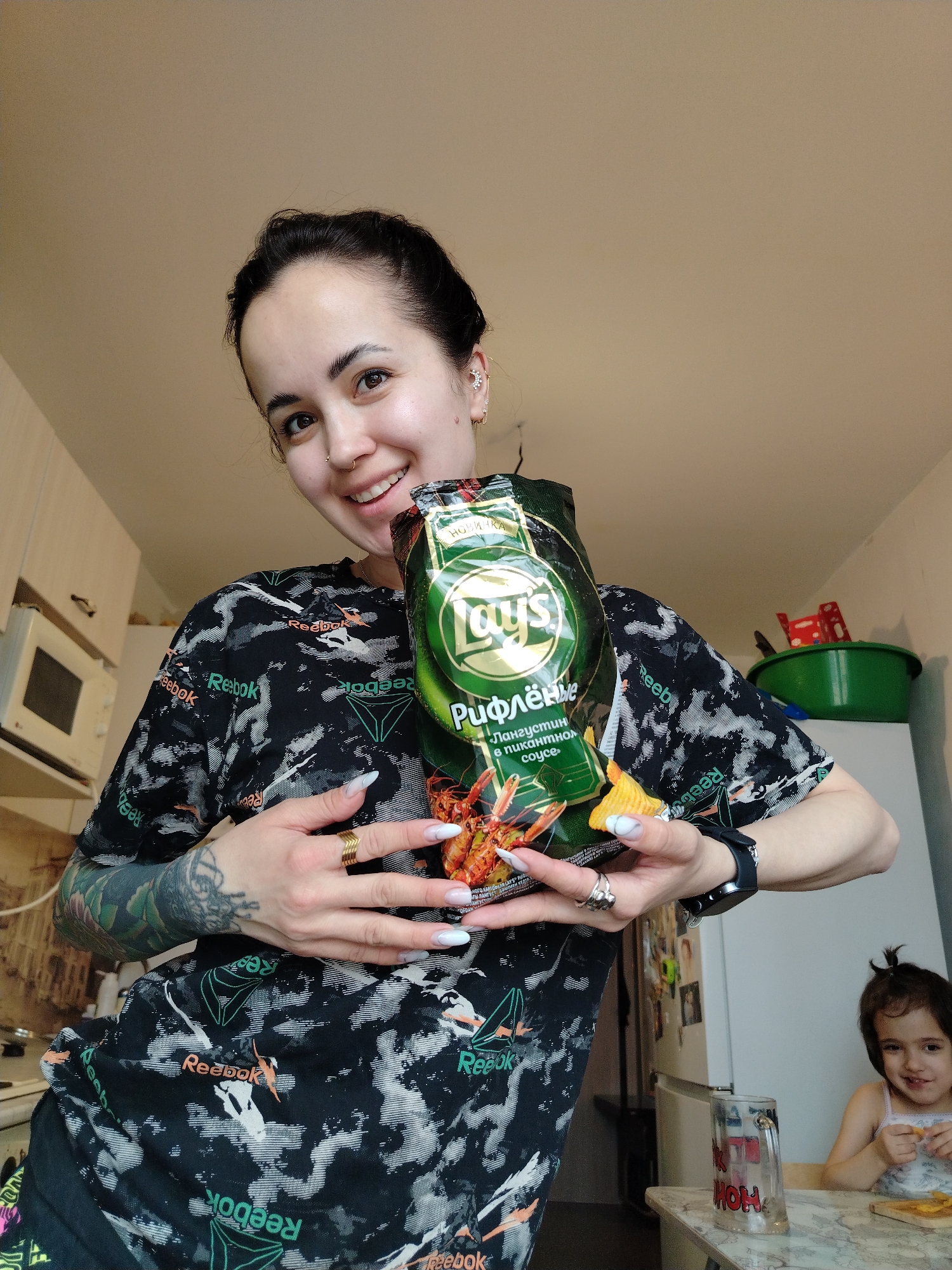 A young woman is indoors, smiling and holding a bag of Lay's Ruffles potato chips with "Langoustine in Spicy Sauce" flavor, written in Russian. She is wearing a black Reebok t-shirt with a white and black pattern and the Reebok logo in green and orange. The woman has a nose ring, multiple earrings, tattoos on her left arm, and rings on her fingers. In the background, there is a young child sitting at a table, also eating chips. A microwave is visible to the left, and a refrigerator is to the right. A green plastic container sits atop the refrigerator. A decorative glass is on the table in front of the child.