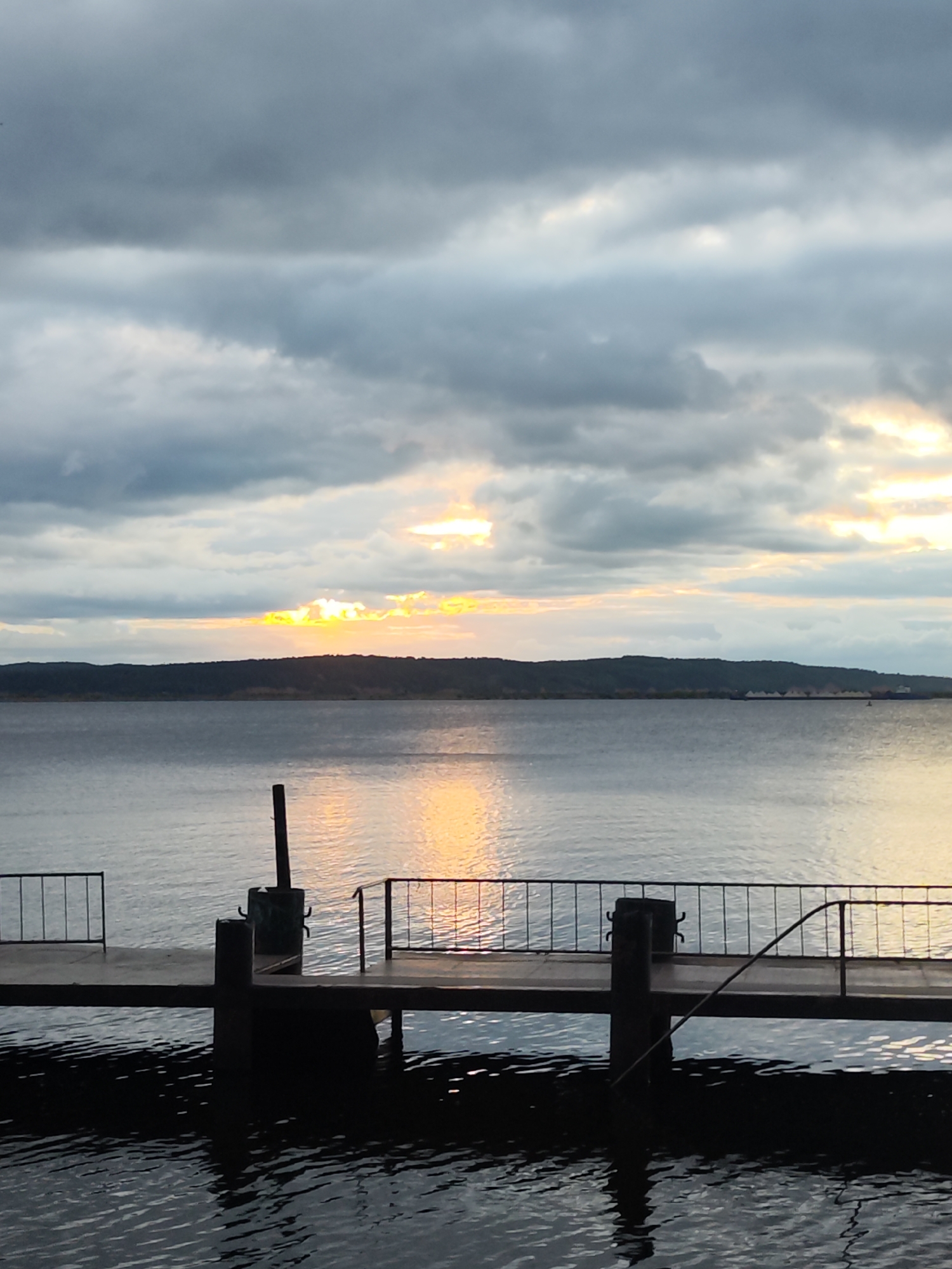A sunset casts a golden glow through dark, cloudy skies over a wide, calm body of water. A dark, tree-lined horizon stretches across the middle ground. In the foreground, a dark wooden dock extends into the water, featuring a railing on either side. The dock's reflection is visible in the rippling water below.