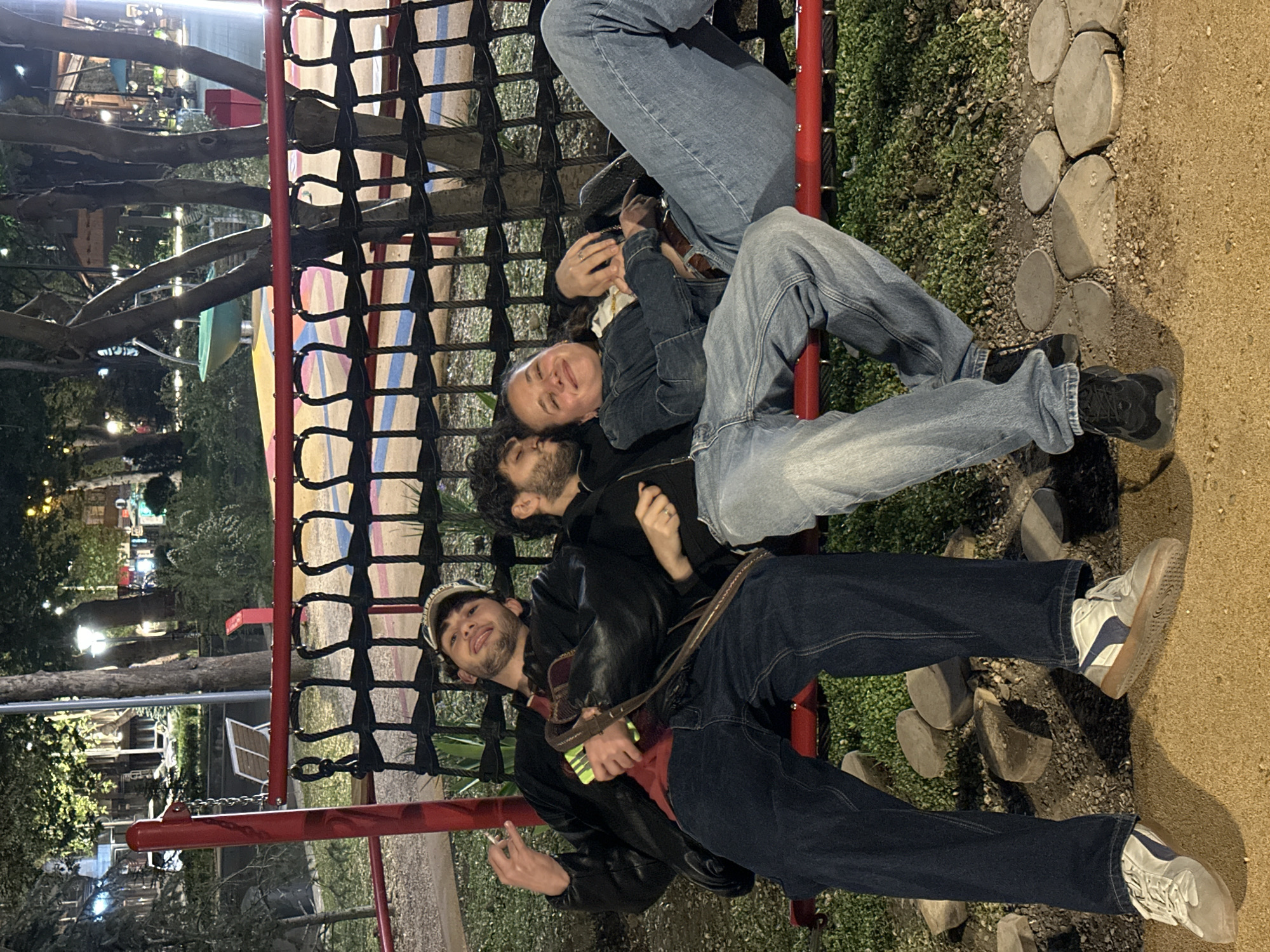Three individuals are situated on a red playground climbing structure at night. The person at the bottom of the image, a young adult with dark hair and a baseball cap, is wearing a black jacket over a red shirt and dark blue jeans. He is holding a small rectangular object and a cigarette in his hand. Above him is a person with a beard and dark hair, wearing a black jacket. Resting above that person is a female with light brown hair wearing a denim jacket and light blue jeans. The location is in Yerevan, Armenia. The environment appears to be a park with trees and lights in the background. A stone walkway is next to the climbing structure.