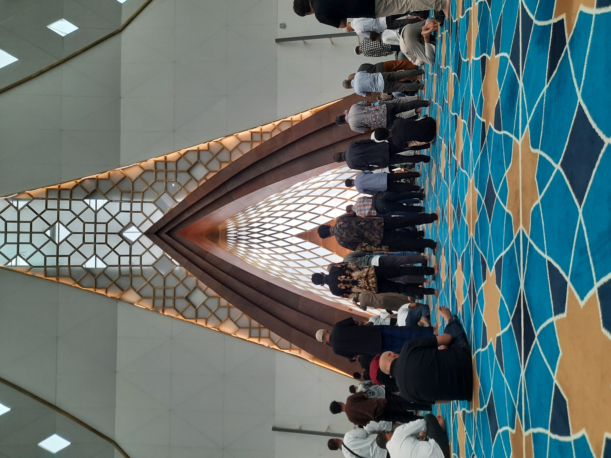 The image depicts a group of people engaged in prayer within a modern mosque. The interior features a striking architectural design with a triangular, vaulted ceiling adorned with a geometric lattice pattern, illuminated by natural light filtering through. The floor is covered with a vibrant blue and gold patterned carpet. Several individuals are seen standing and kneeling in prayer, facing the architectural focal point. The scene suggests a communal religious gathering.