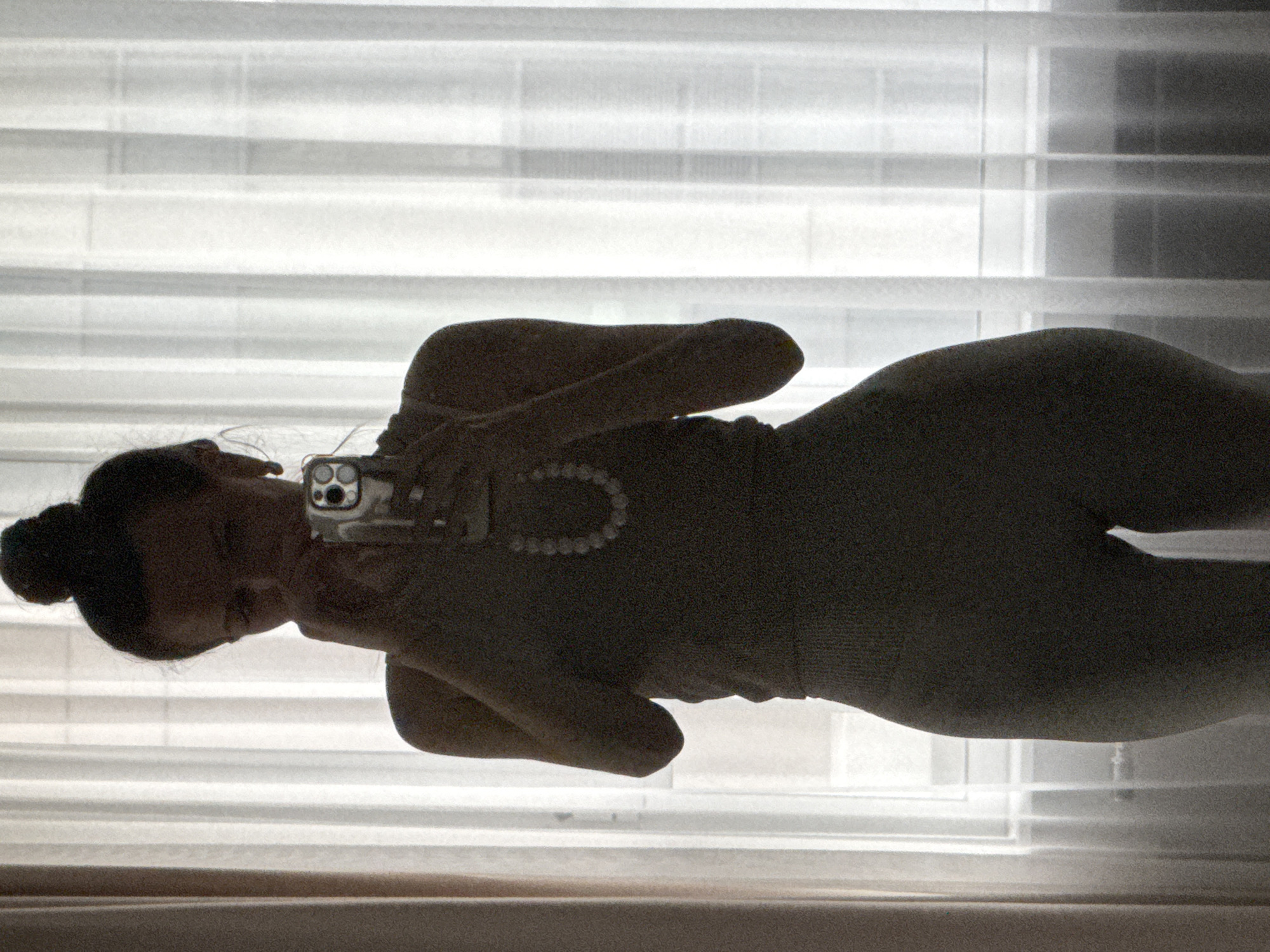 The original image is presented rotated 90 degrees clockwise. It features a person, likely female, captured in a strong silhouette against a brightly lit window with horizontal blinds, suggesting an indoor setting in Khimki, Russia. The person is positioned horizontally, with their head to the left side of the frame, and is shown from approximately the head to mid-thigh.
The primary activity depicted is the person holding a smartphone to take a selfie or record themselves. The phone, held in their right hand, is visible with its multiple camera lenses facing outwards. A beaded bracelet or necklace is visible around their left wrist or arm. Their hair is neatly tied back in a bun.
The lighting is characterized by strong backlighting from the window, which casts the person's figure into deep shadow, creating a prominent silhouette. This makes specific facial features, clothing details, and emotional expressions indistinguishable. The bright background suggests it is daytime. The image has a noticeable grainy texture. No discernible text is visible within the frame.