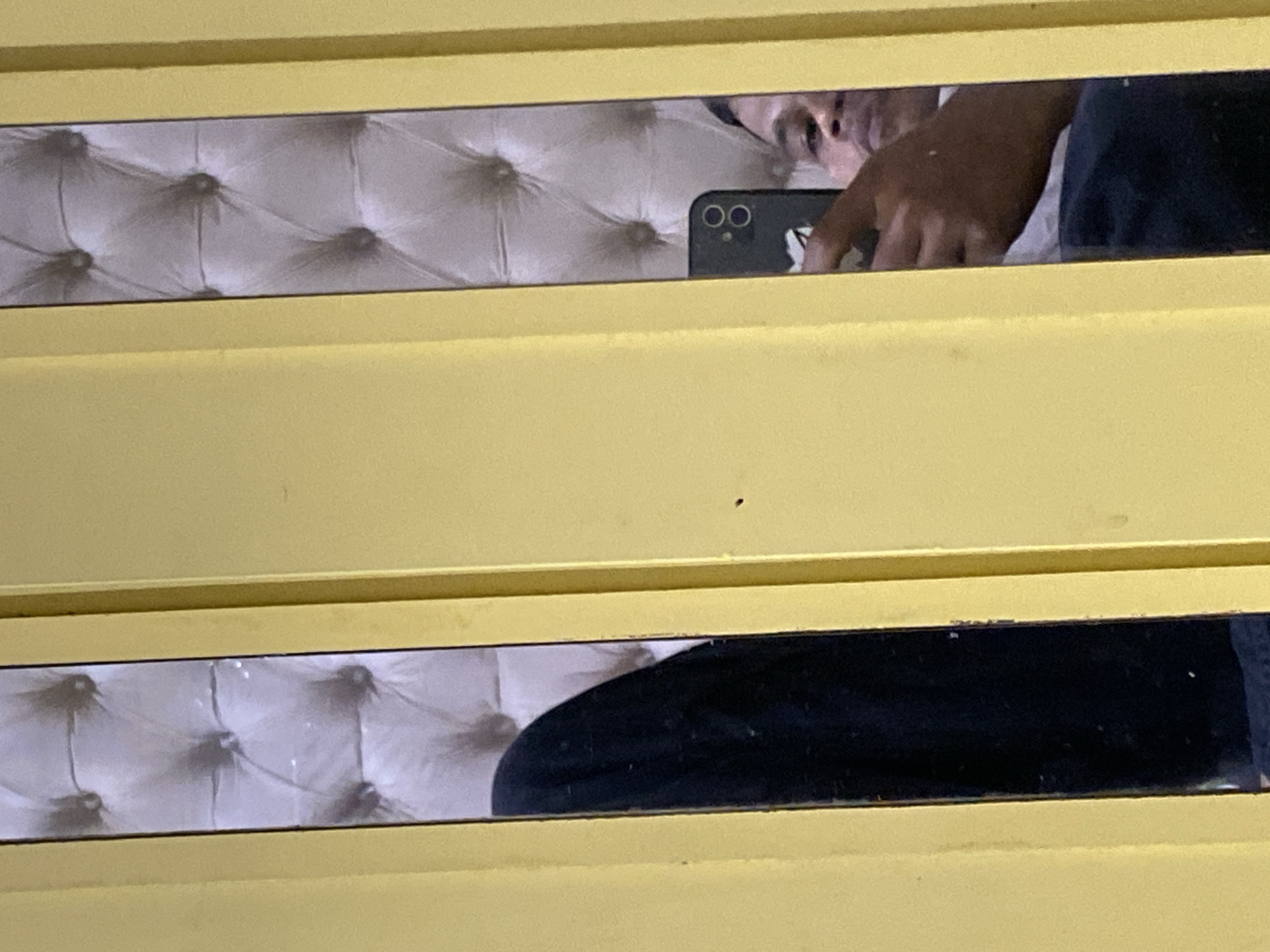 In the city of Unknown, Unknown, a person is taking a mirror selfie. The person is holding a smartphone and their face and hand are visible in the reflection. The background of the mirror shows a tufted headboard, likely from a bed, and a dark fabric, possibly clothing. The scene is framed by a yellow structure, which could be a doorway or a piece of furniture.