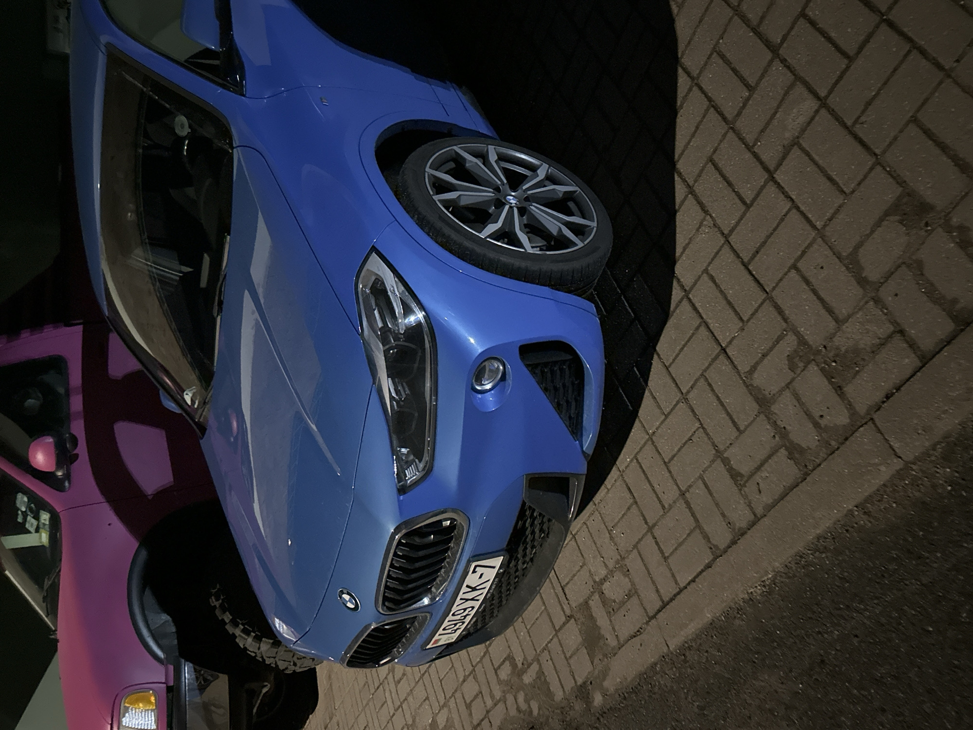 The image shows a blue BMW car parked on a brick-paved surface, likely a sidewalk or driveway. The car's license plate reads "L-X-6767". Next to the BMW is a pink vehicle, possibly a pickup truck, partially visible. The setting is at night or in low light, with artificial lighting illuminating the vehicles. The brick pattern of the paving suggests an urban or suburban location. The license plate indicates the car is likely registered in Belarus, and based on that and the location of the license plate provider mentioned, we can speculate that this image was taken in Minsk, Belarus.