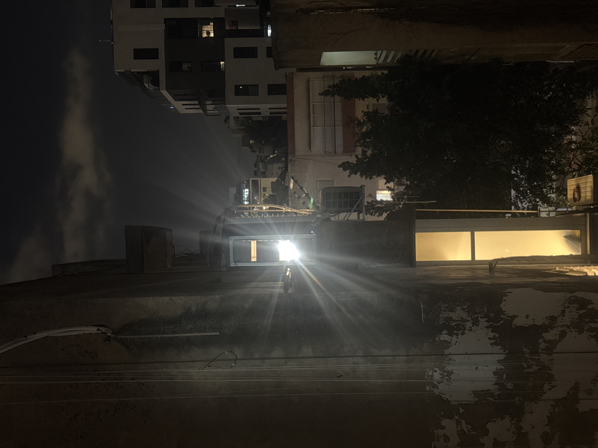The image shows a night view in Ramat Gan, Israel. The foreground is dominated by a flat, weathered rooftop with visible wiring and peeling paint. A bright light source, likely a window, shines intensely from a building behind the rooftop, creating strong light rays. Further back, multi-story buildings are visible, with some windows illuminated. To the right of the central building, a tree is present, and a streetlight fixture is affixed to the side of the building. A cloud formation is visible in the dark sky to the left.
