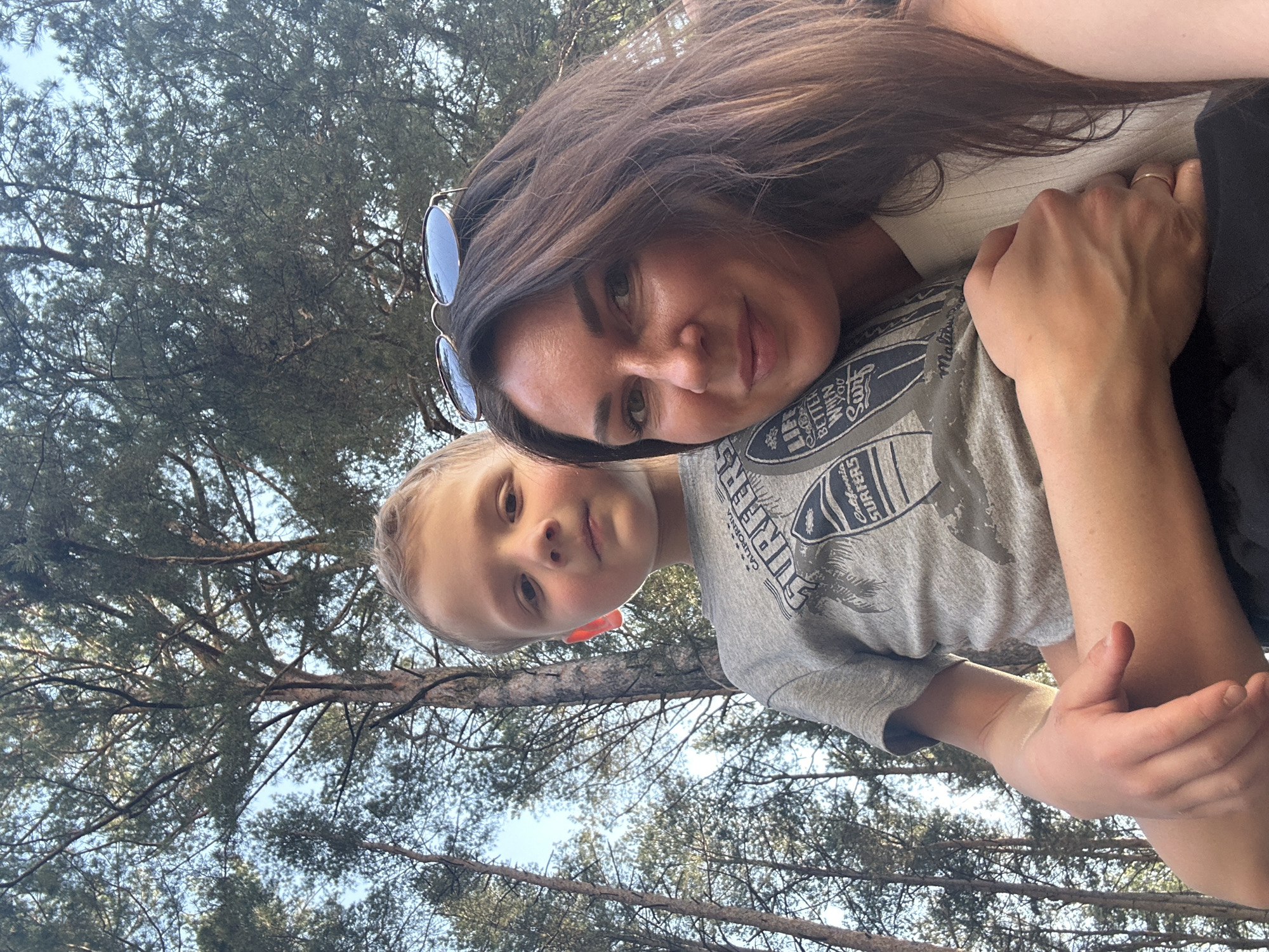 A woman and a child are photographed outdoors in a wooded area. The woman has long dark hair and sunglasses perched on top of her head. She is wearing a light-colored top. The child, a young boy, has light blonde hair. He is wearing a grey t-shirt with surf-themed graphics. The woman is holding the child close. In the background, there are trees with dense foliage and some patches of blue sky visible through the branches. The location is likely in Chaikovsky, Russia, as requested.
