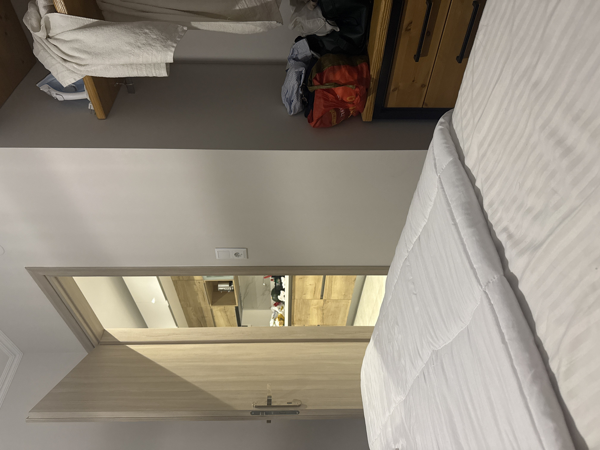 The image depicts an interior scene, possibly a bedroom in Ποτός, Greece, with a view into a kitchenette. In the foreground, a neatly made bed with white striped sheets and a white comforter occupies the lower half of the frame. To the right of the bed, a wooden dresser with black handles is partially visible. Above the dresser, an open closet reveals a shelf holding an iron and folded white towels. Below the shelf, various bags and folded clothes are stored.
The doorway on the left is ajar, revealing a glimpse of a modern kitchenette. The kitchenette features wooden cabinetry, a white countertop with items on it, and a tiled backsplash. The walls are painted a neutral, light color. The overall impression is one of a clean and functional living space.