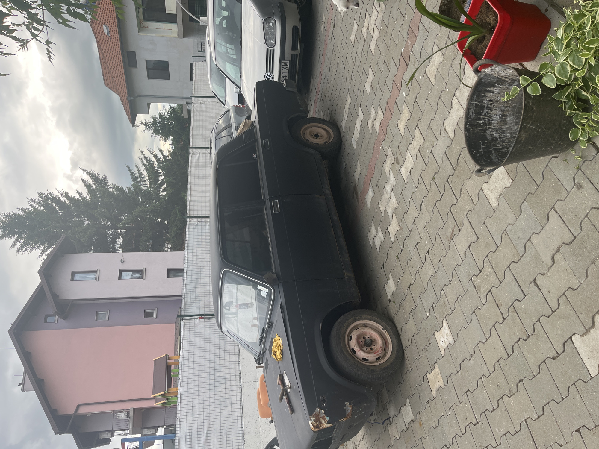 The image depicts an outdoor scene in Podgumer, Bulgaria, with a focus on several parked vehicles and residential buildings. In the foreground, a weathered black sedan, appearing to be a Lada, is parked on a cobblestone driveway. The car shows signs of disrepair, with rust visible on the front bumper and fender. Tools, including a hammer and a cloth, are placed on its hood. To the right of the Lada, a silver Volkswagen Golf wagon is parked. A small white dog is visible in the lower right corner, looking towards the camera.
Behind the cars, a white construction fence runs across the frame, separating the driveway from the background. Beyond the fence, two residential buildings are visible. The building on the left is a multi-story structure with a pink and light purple facade, featuring balconies and windows. A small playground structure is also visible near this building. To the right, another residential building with a red tile roof and white walls is under construction or renovation, with visible scaffolding and unfinished sections. Evergreen trees are present in the background, and the sky is overcast with clouds. The overall impression is that of a residential area with ongoing construction and a mix of older and newer vehicles.