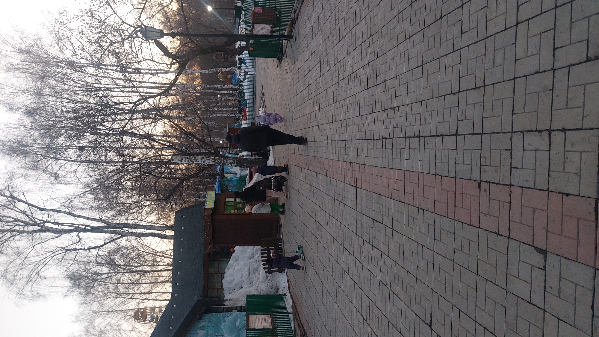 The image depicts a section of the Novosibirsk Zoo in Novosibirsk, Russia. The scene is set on a paved walkway, constructed of gray and red bricks. Along the walkway, a group of people, including adults and children, are positioned near what appears to be a small kiosk or shop with a dark brown facade. A pile of snow is located adjacent to the kiosk, indicating recent or lingering winter conditions.
Bare trees, characteristic of the late fall or winter season, frame the view in the background and on the left side of the image. A vintage-style street lamp is also visible. In the distance, what seems to be a carousel or another ride is discernible. One person is walking along the walkway.