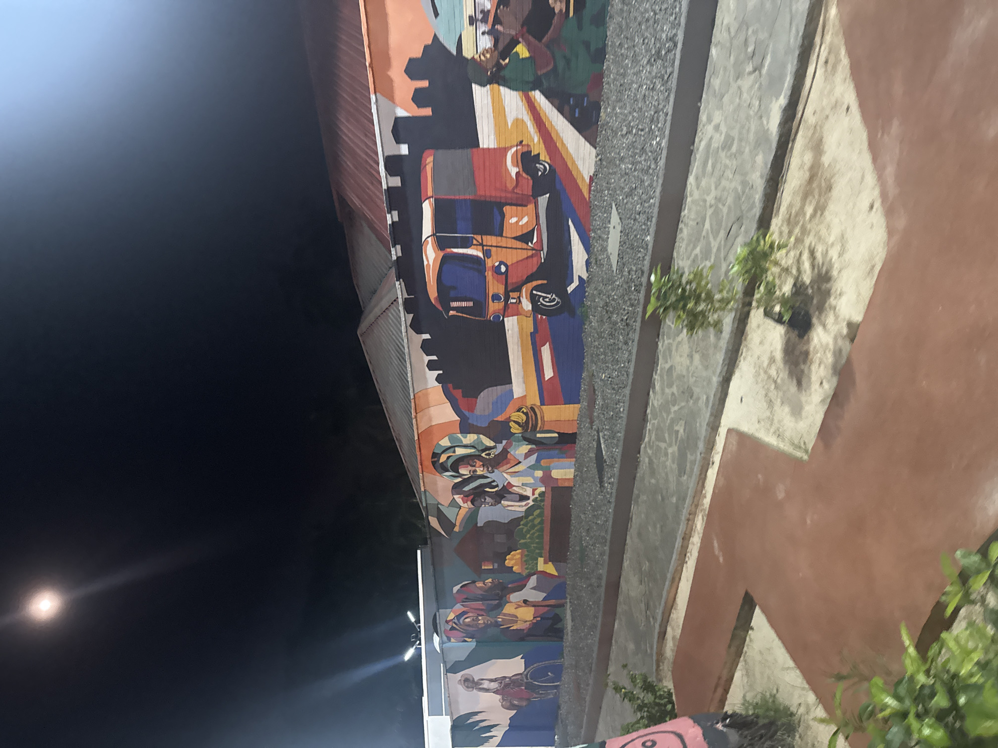 The image captures a vibrant street art mural on the side of a building at night, likely in Uyo, Nigeria, given the context. The scene is illuminated by artificial lights, with bright flares visible against the dark sky.
The mural itself is rich with depictions of local life and culture. In the upper section, a stylized cityscape with dark building silhouettes forms a backdrop. Below this, a prominent orange and blue auto-rickshaw, commonly known as a "keke napep" in Nigeria, is painted, suggesting bustling urban transport.
Further down the mural, several figures are depicted. Many wear colorful headwraps and traditional attire, engaging in what appears to be market activities or social gatherings. Baskets filled with produce, possibly fruits or vegetables, are visible amongst these figures. Towards the bottom, the mural continues to portray people, including one figure playing a trumpet and another seated in a wheelchair, suggesting inclusivity and diverse community representation. The art style is bold and colorful, using strong lines and geometric shapes to create a dynamic and energetic composition.
Adjacent to the mural, a paved pathway or ground area runs alongside the building, featuring textured concrete and earthy-toned sections, with small green plants growing intermittently. The building itself has a corrugated metal roof visible at the top edge of the image. The overall impression is a lively public space adorned with expressive art, celebrating local life and culture under the night sky.