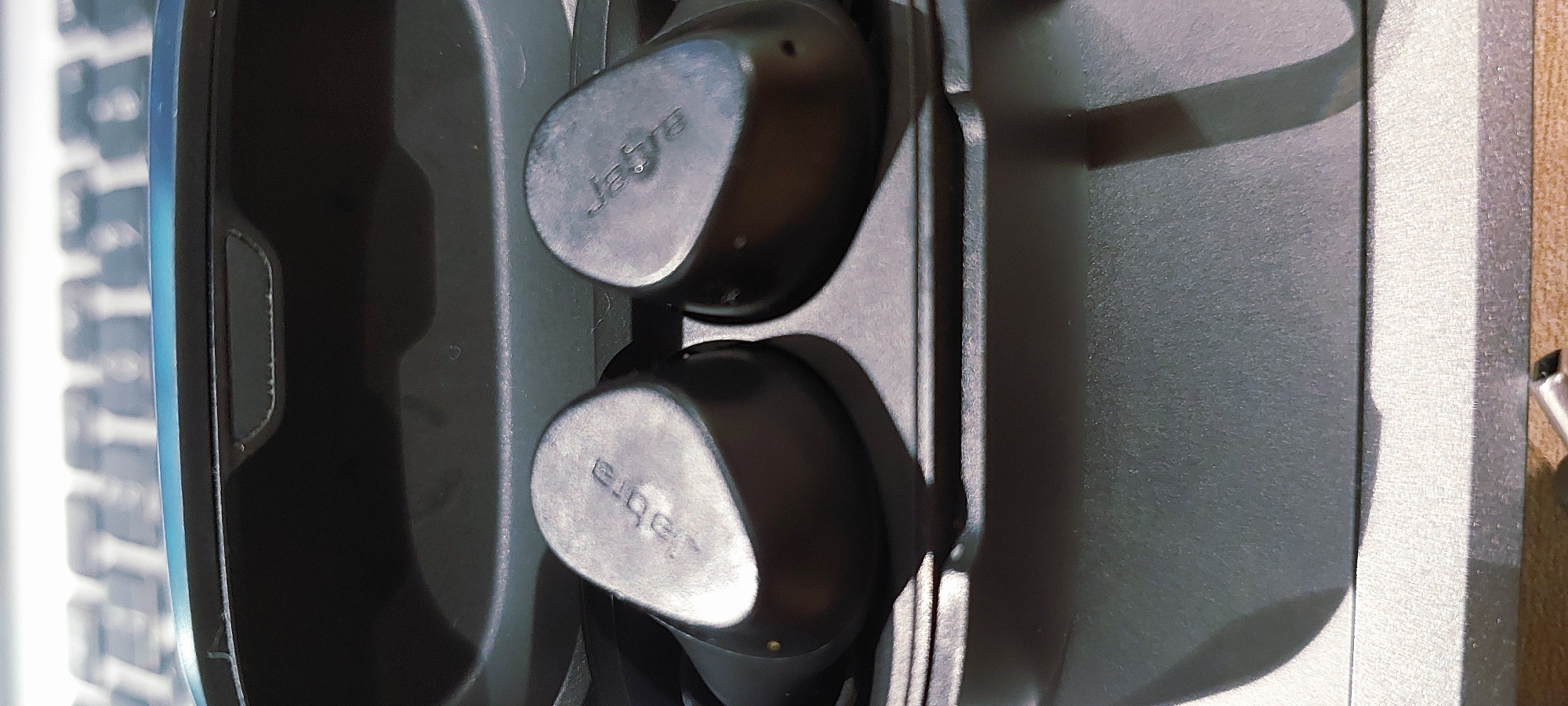 The image shows a pair of black Jabra wireless earbuds in their charging case. The earbuds are positioned side-by-side, with the Jabra logo visible on each. The charging case is dark gray and appears to be open, revealing the indentations where the earbuds sit. The background is out of focus, but suggests a keyboard-like surface with a bluish tint. The overall lighting is subdued, with shadows cast on the charging case. There are no people or animals present in the image, and no signs of the location in Pagedangan, Indonesia.