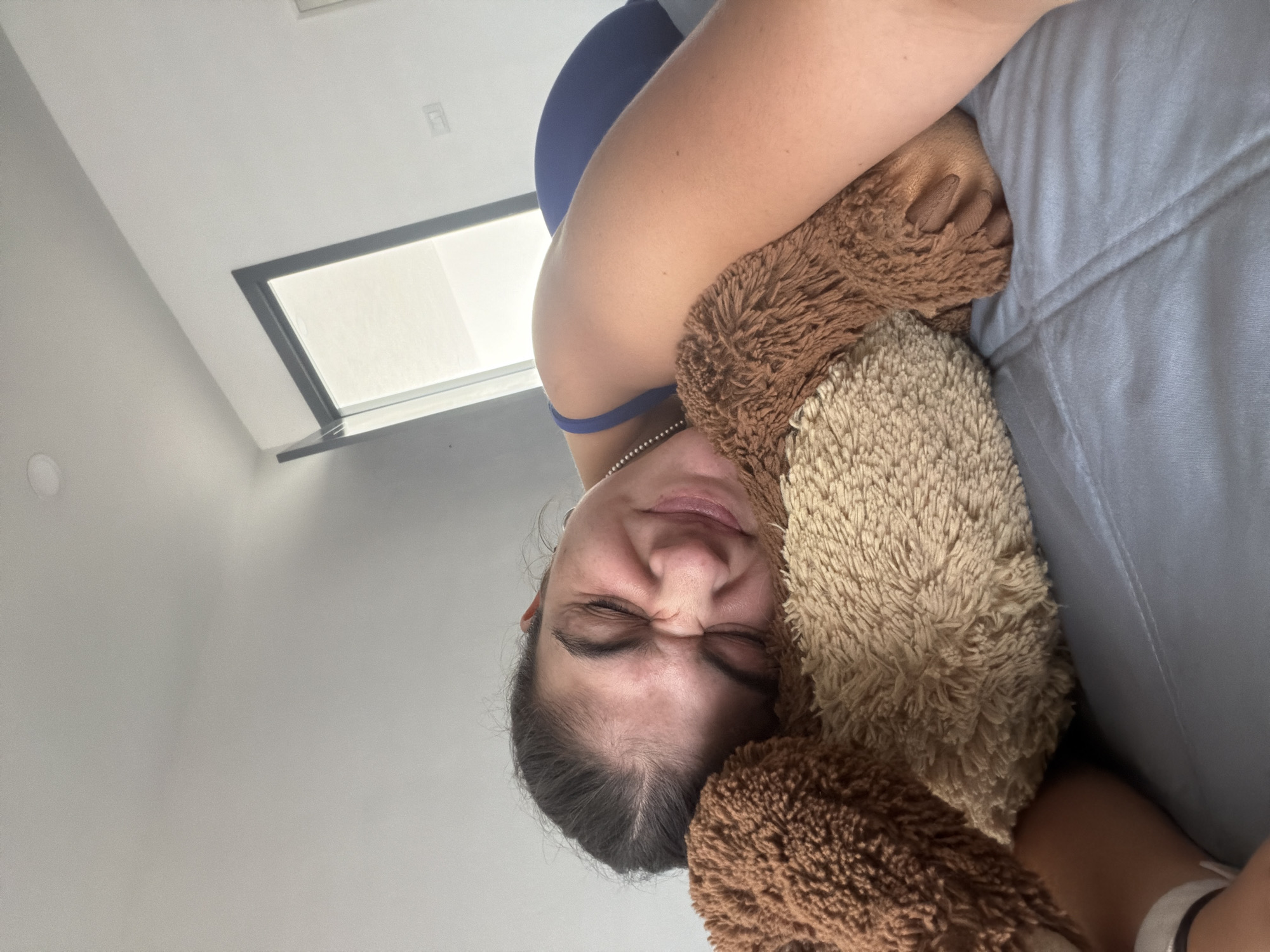 A woman with dark hair and closed eyes is holding a stuffed bear. She is wearing a blue top and is positioned next to a gray sofa. A window is visible in the background. The photo appears to have been taken indoors. It is not possible to determine the location in the city of Puebla, Mexico from this image.