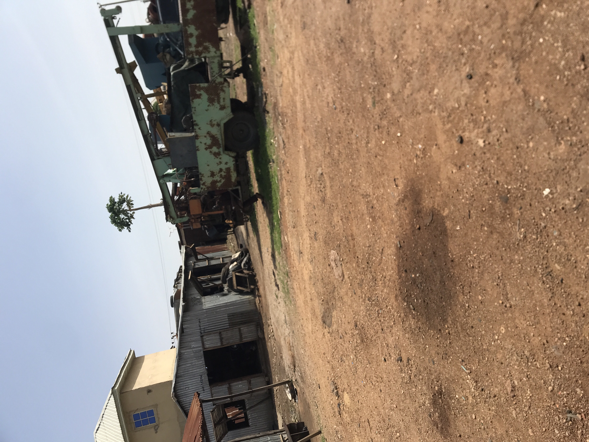 This media file presents an outdoor scene in what appears to be an industrial or workshop area in Rafin Par, Nigeria, captured on a bright, clear day. The image is vertically oriented but depicts a horizontal landscape.
In the midground, a large, significantly rusted, light-green piece of heavy machinery, possibly a forklift or a component of a crane, stands prominently on reddish-brown dirt. A single black wheel is visible on this machine. Patches of green grass are scattered around the base of the equipment. The ground is unpaved, consisting of dry dirt mixed with small stones and some minor debris. A distinct shadow is cast on the ground, suggesting bright sunlight.
To the left of the frame, there is a light-colored building, possibly cream or light yellow, featuring a clear blue-paned window. Adjacent to this building is another structure made of corrugated metal, likely a workshop or storage shed, with a dark, open doorway. In the upper portion of the image, against a clear, light blue sky, a small, sparse tree with green leaves is visible.
No people are present in the image, and there is no explicit activity taking place. The clear sky and strong lighting indicate it is daytime. No visible text is discernible on any of the structures or objects.