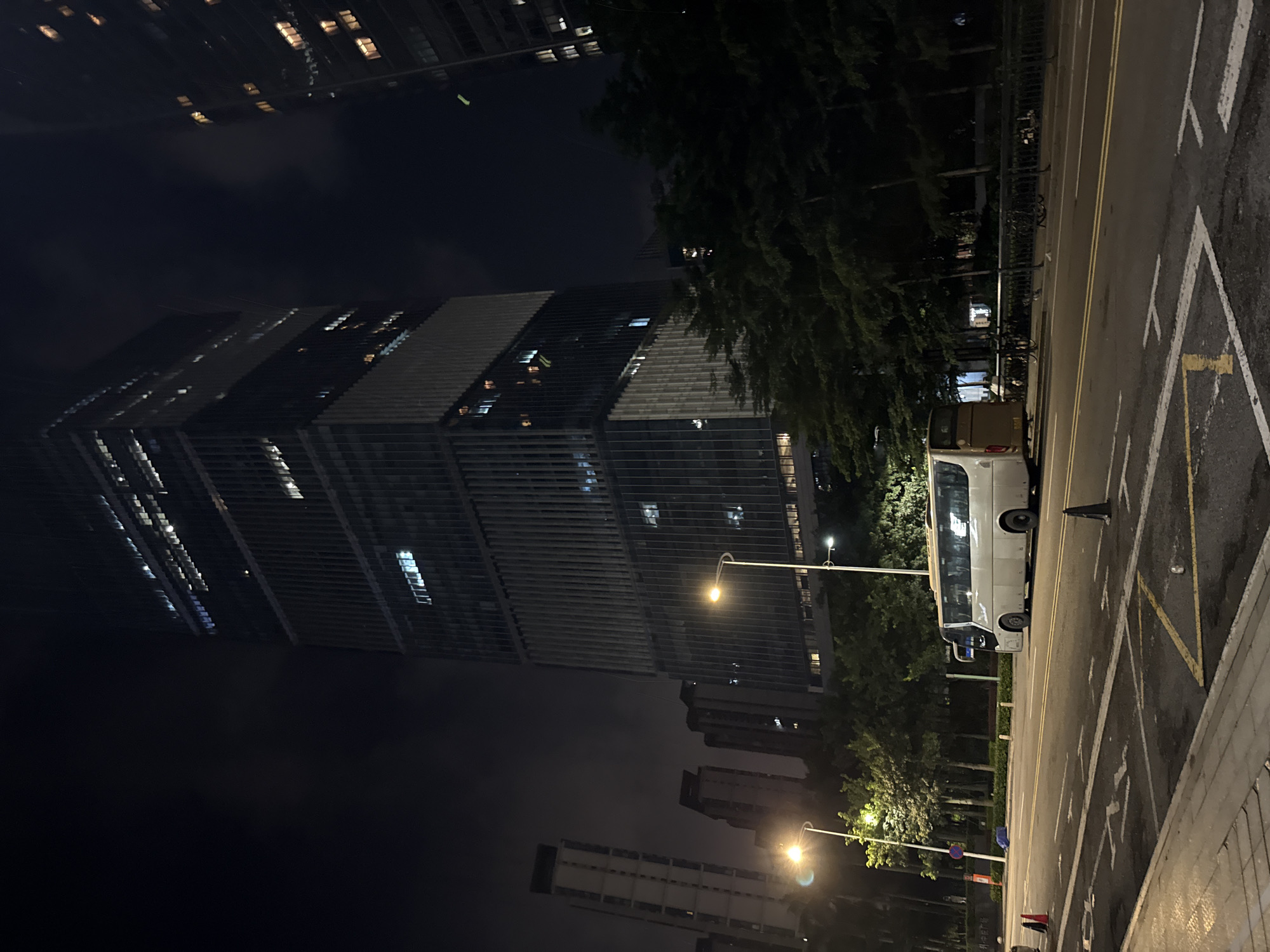 This media file depicts a nighttime urban scene in Haizhu District, China. The primary subject is a tall, modern skyscraper with numerous illuminated windows, suggesting it is occupied. In front of the skyscraper, trees with dense foliage line a street. A white bus is parked on the side of the road, alongside a curb marked with yellow and white lines, indicating a parking area or a designated zone. Streetlights illuminate the scene, casting a warm glow on the asphalt and the surrounding environment. The sky is dark, typical of nighttime, and there are no indications of specific weather conditions. The overall mood is one of quiet urban activity after dark, with the illuminated buildings and streetlights creating a sense of a functioning city at night. There are no visible people or explicit text in the image. The image is captured from a street-level perspective, looking upwards towards the skyscraper and across the street.