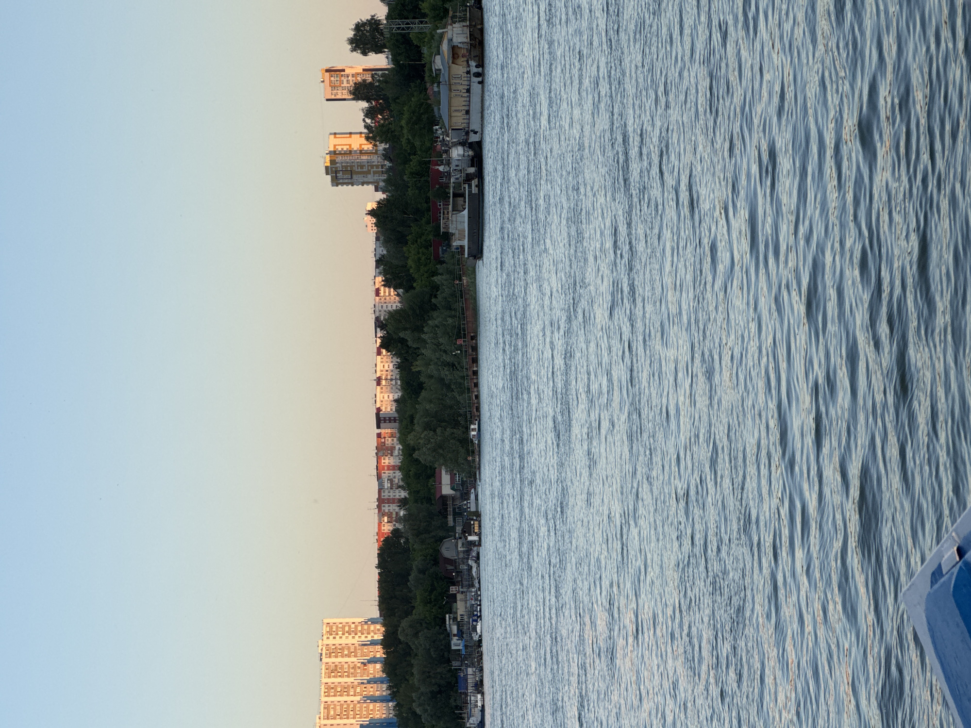 The image depicts a wide river in Novosibirsk, Russia, with a cityscape in the background. In the foreground, the river's surface is rippled, reflecting the light of what appears to be late afternoon or early evening sun. The color of the water is a deep blue, with lighter highlights on the crests of the waves.
The middle ground shows a shoreline lined with dense green trees. Along the water's edge, there are several boats and structures, including what appear to be small buildings and docks. A longer, flat-topped boat or barge is moored near the shore, with a yellow building attached to it. Another, darker vessel is alongside it. Further along the bank, more boats of various sizes are visible, some of which appear to be leisure craft.
The background is dominated by the Novosibirsk skyline. On the left side, a large, pale orange apartment building stands tall, its numerous windows catching the sunlight. To its right, a line of residential buildings stretches across the horizon, some with red roofs. In the center-right of the frame, two modern, multi-story buildings with prominent balconies or architectural features are visible. These buildings have a mix of yellow, white, and orange facades. The sky above is a clear, soft blue, transitioning to a pale yellow or peach hue near the horizon, indicating a setting sun. The overall impression is a view of an urban area situated along a major river, likely during a pleasant time of day.