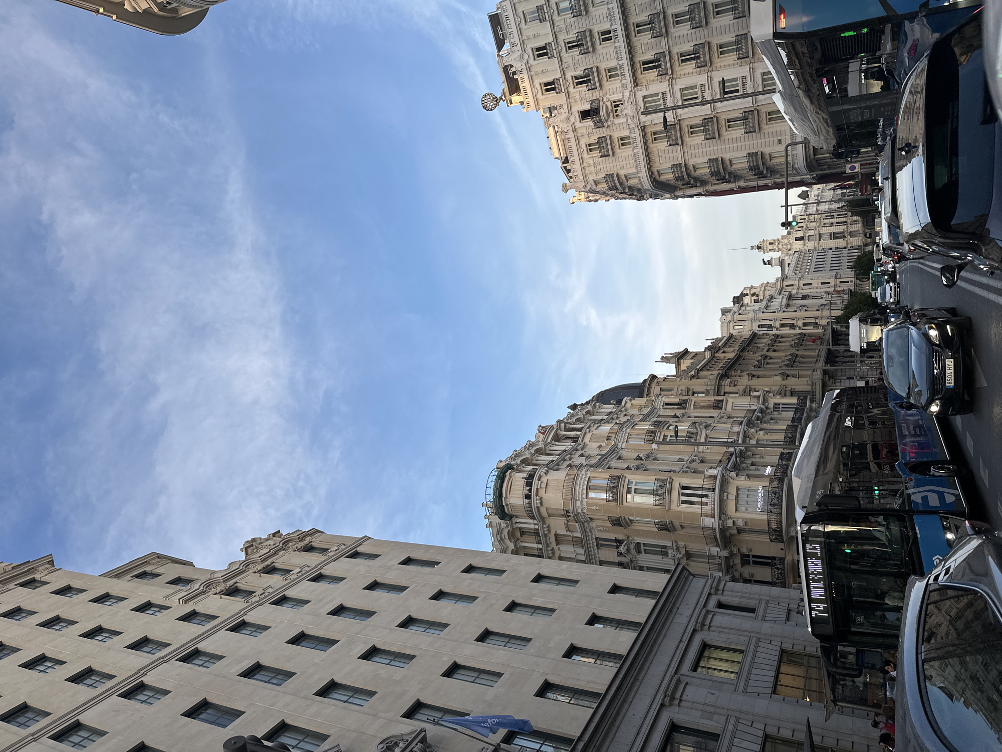 This photograph depicts a street scene in Madrid, Spain, characterized by tall buildings and vehicular traffic. The image is taken from a low angle, looking upwards towards the sky. The sky is predominantly blue with wispy clouds. Several buildings, some with ornate facades, line the street. A bus and multiple cars are visible on the right side of the image, suggesting the scene takes place on a busy street. The presence of buses and cars implies that the location is an urban environment. The style of architecture indicates the location is Madrid.
