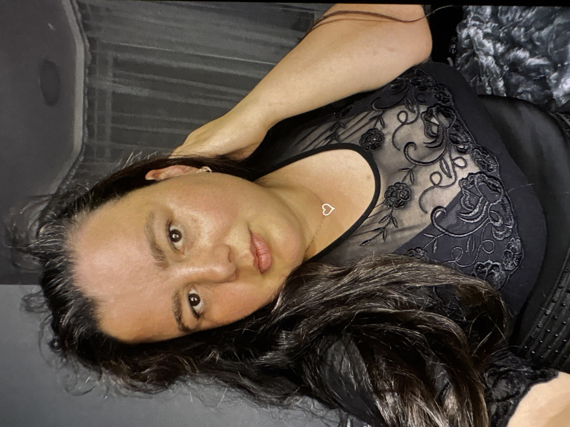A woman is pictured in a close-up, waist-up shot, looking at the camera. She has long, dark, wavy hair that falls over her left shoulder and a portion of her chest. She is wearing a black, sheer top with floral embroidery over a black undergarment. A silver necklace with a heart-shaped pendant rests on her chest. Her right hand is raised and rests near her head, with her fingers touching her hair. She is wearing small, hoop earrings. The background is dimly lit and appears to be an indoor setting, possibly a bedroom, with sheer curtains visible behind her. The overall lighting is soft and natural, with some shadows on her face. The image is a selfie, taken from a slightly high angle.