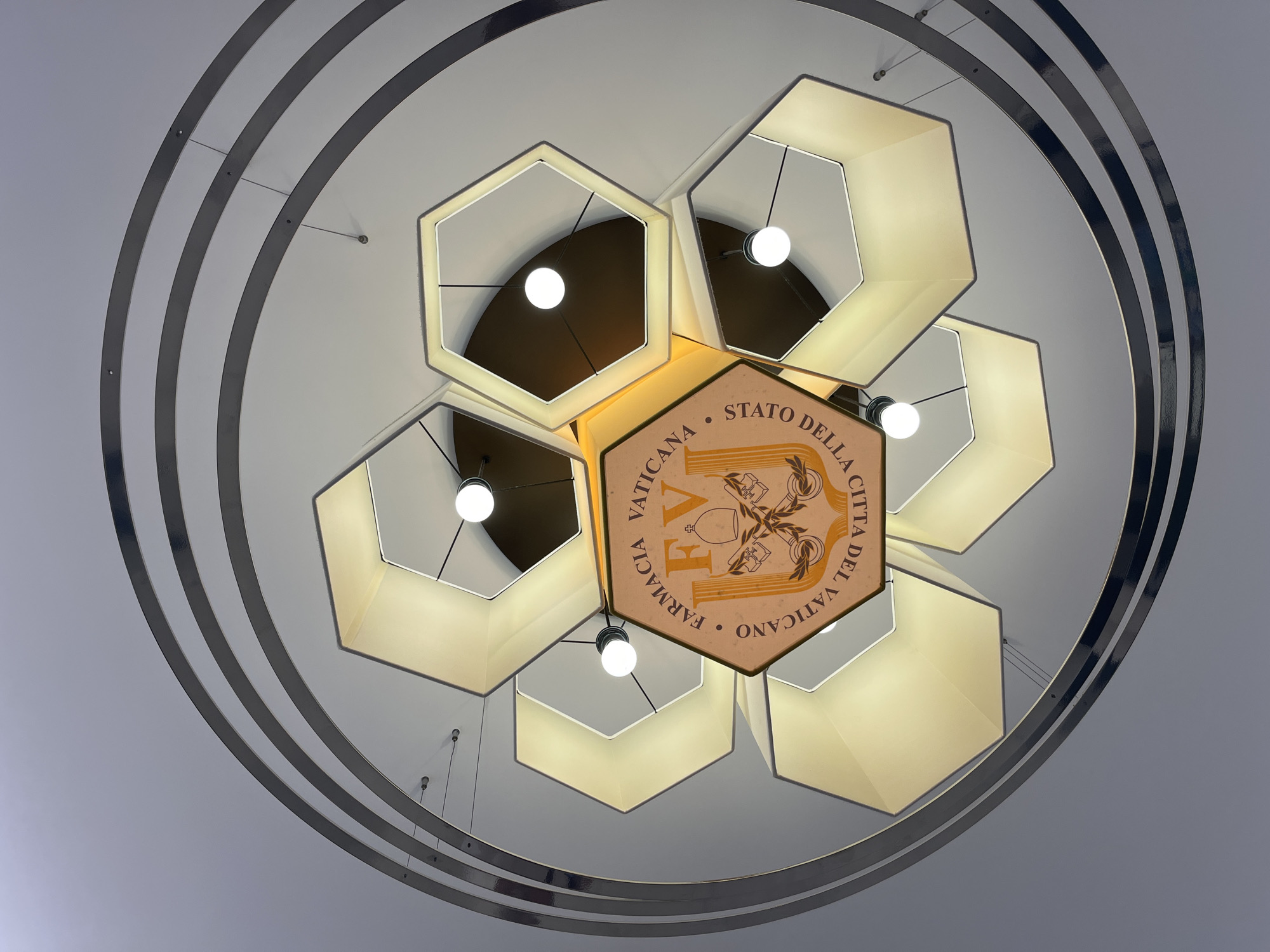 This image shows a downward view of a hexagonal chandelier in Città del Vaticano, Vatican City. The chandelier has a central hexagonal panel featuring the text "FARMACIA VATICANA" and "STATO DELLA CITTA DEL VATICANO" along with the initials "FV" and an emblem of crossed keys and a miter. Surrounding the central panel are six other hexagonal light fixtures, each with a bulb illuminating the interior. The entire structure is suspended by thin wires from a ceiling, and is framed by three concentric, curved metallic rings. The lighting is warm and ambient. There are no people or animals visible in the image.