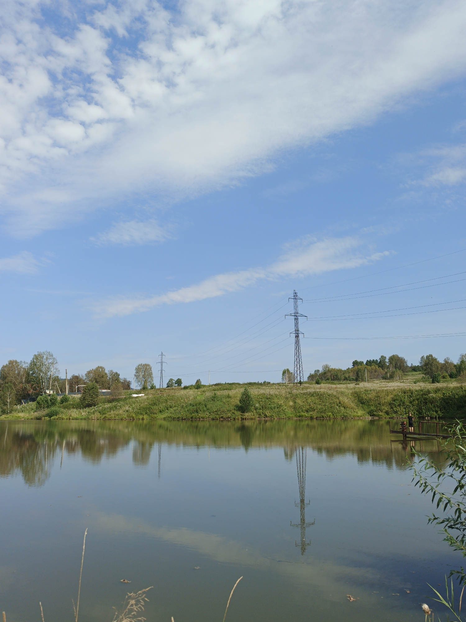 The image depicts a serene lake scene in the city of Urtiushka, Russia. The water surface reflects the clear blue sky, dotted with wispy white clouds. The foreground shows the calm water with some reeds and fallen leaves. Across the lake, a grassy embankment is covered with lush green vegetation and scattered trees. A prominent feature on the embankment is a tall, dark electrical pylon with wires extending across the horizon. In the distance, a few houses and structures can be seen nestled amongst the trees. To the right of the image, a small wooden platform extends into the water, where a lone figure, a person wearing dark clothing, stands. The overall impression is one of peaceful rural tranquility.