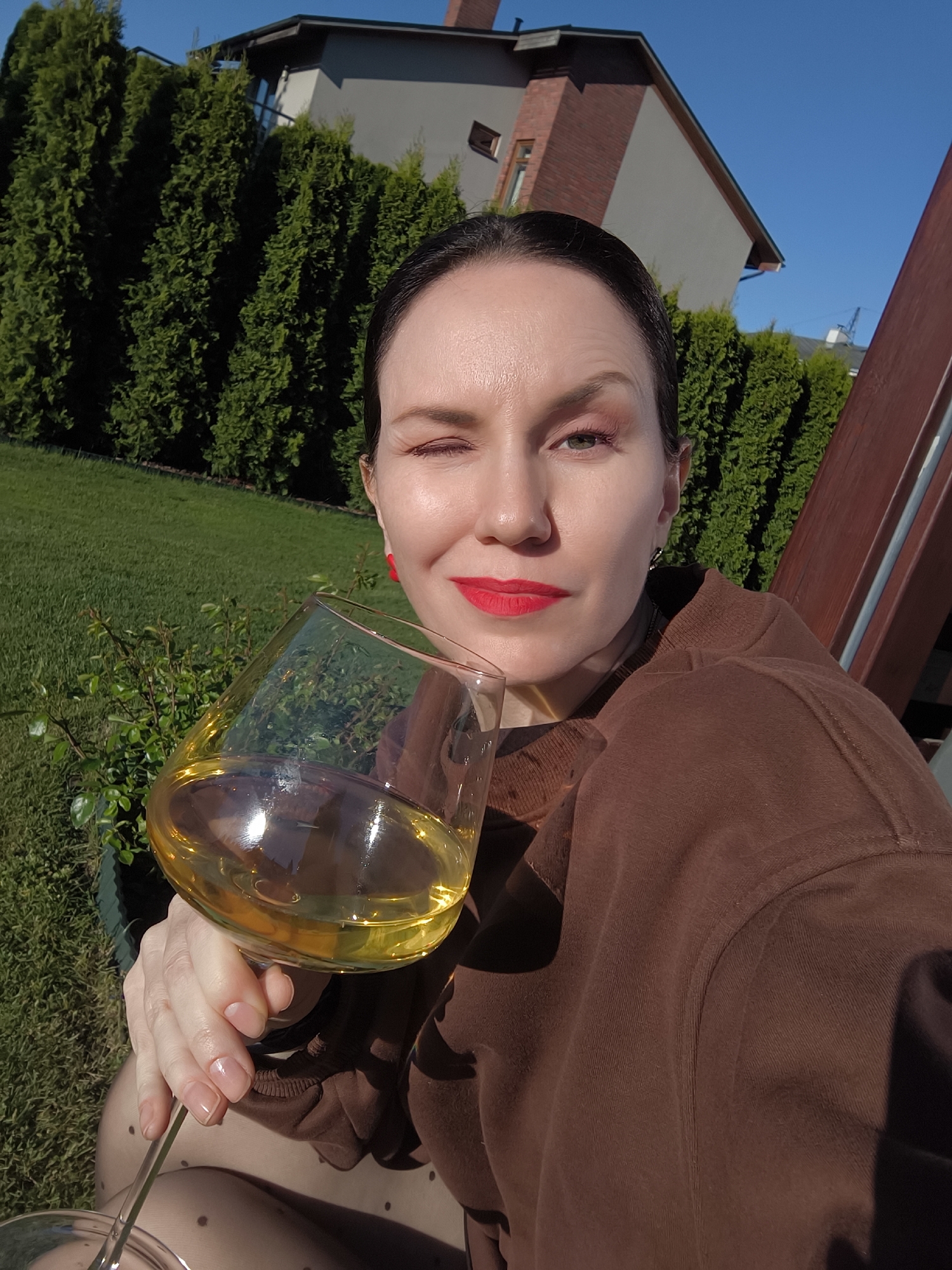 The image shows a woman holding a large glass of a light-colored liquid, possibly white wine, while winking at the camera. She has dark hair pulled back, fair skin, and is wearing red lipstick. She is dressed in a brown hooded sweatshirt. Red earrings are visible on her left ear.
The background consists of a green lawn, tall green hedges or trees, and a modern house with grey and brick elements under a clear blue sky. The woman is sitting on what appears to be a wooden chair. The location is not discernible from the image.