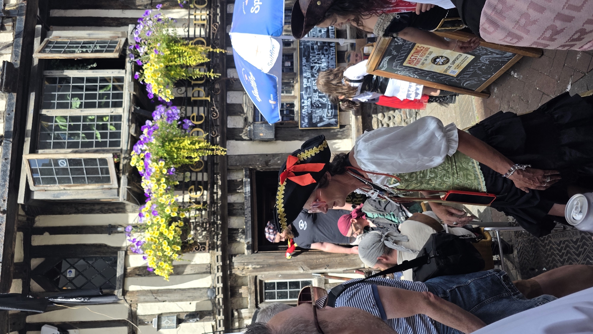 The image depicts a street scene in St Leonards-on-Sea, United Kingdom, possibly during a pirate-themed event. In the foreground, a woman dressed in a pirate costume, complete with a tricorn hat adorned with a red ribbon, is the central focus. She is wearing a white blouse with puffed sleeves and a green corset over a long, black ruffled skirt. A red mobile phone is attached to her chest by a lanyard. Her hands are visible, adorned with several rings and a bracelet.
Behind her, to the left, a man with a beard, wearing a black t-shirt, stands near the entrance of a building. He has a red parrot accessory attached to his shirt. Other individuals in pirate attire are partially visible, including a woman to the right wearing a dark hat and a black outfit.
The background features a historic building with timber framing and multiple windows, some of which are open and decorated with hanging baskets filled with yellow and purple flowers. A blue and white umbrella is set up to the right of the entrance. A chalkboard sign is propped up on an easel, displaying text that includes "HASTINGS PIRATE FESTIVAL JULY 18-19 2016". The ground is paved with cobblestones. The overall atmosphere suggests a festive outdoor gathering.