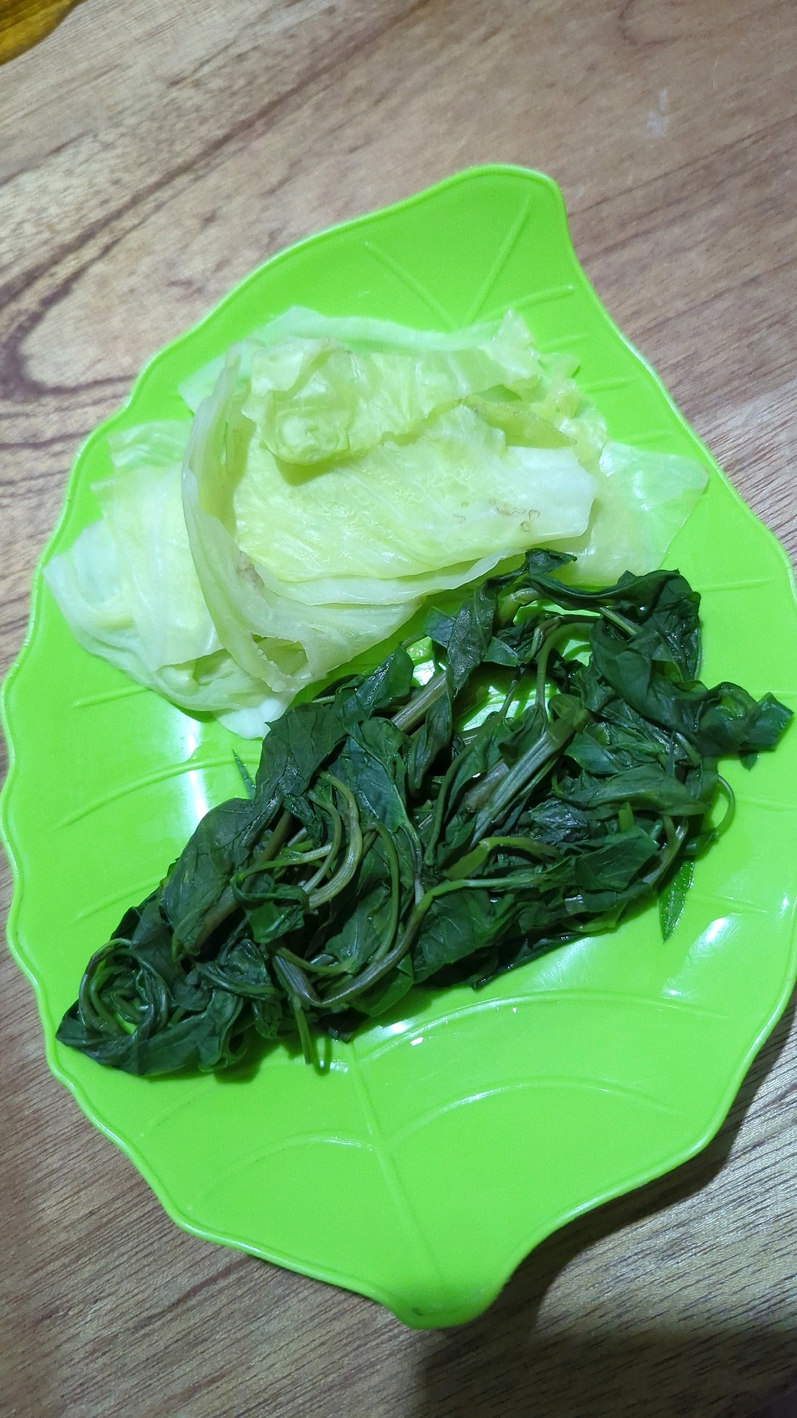 A close-up, high-angle shot captures a bright green, leaf-shaped plastic plate filled with blanched vegetables. The plate rests on a textured wooden surface, likely a table. On the left side of the plate are several pale green, wilted lettuce leaves. To the right, a generous portion of cooked dark green leafy vegetables, possibly spinach or water spinach, is piled. The vegetables appear moist and slightly glossy, suggesting they have been recently cooked. The lighting in the image is somewhat diffused, casting subtle shadows. The scene is simple and domestic, suggesting a home-cooked meal in Baroqah, Indonesia.