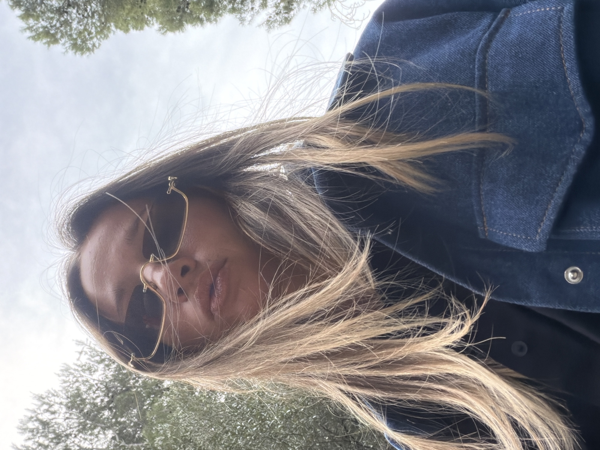 A low-angle outdoor shot depicts a person with blonde hair wearing sunglasses and a denim jacket. The setting appears to be a park with trees visible in the background. The city is Unknown, Unknown. The person is looking down at the camera. The sky is overcast.